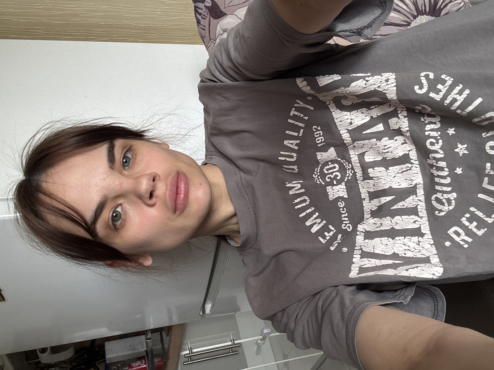 A person with light skin, brown hair, and blue-green eyes is positioned in front of what appears to be a kitchen. The person is wearing a gray long-sleeved t-shirt with a graphic print in white. The shirt reads "VINTAGE Authentic RELIABLES CLOTHES PREMIUM QUALITY Since 1992 -30". The person is looking directly at the camera with a slight pucker of the lips. The background includes a white cabinet and what seems to be a part of a kitchen counter with stainless steel hardware. The scene is indoors, and the lighting appears to be natural. There is no definitive evidence to place this image in the city of Rostov-on-Don, Russia.