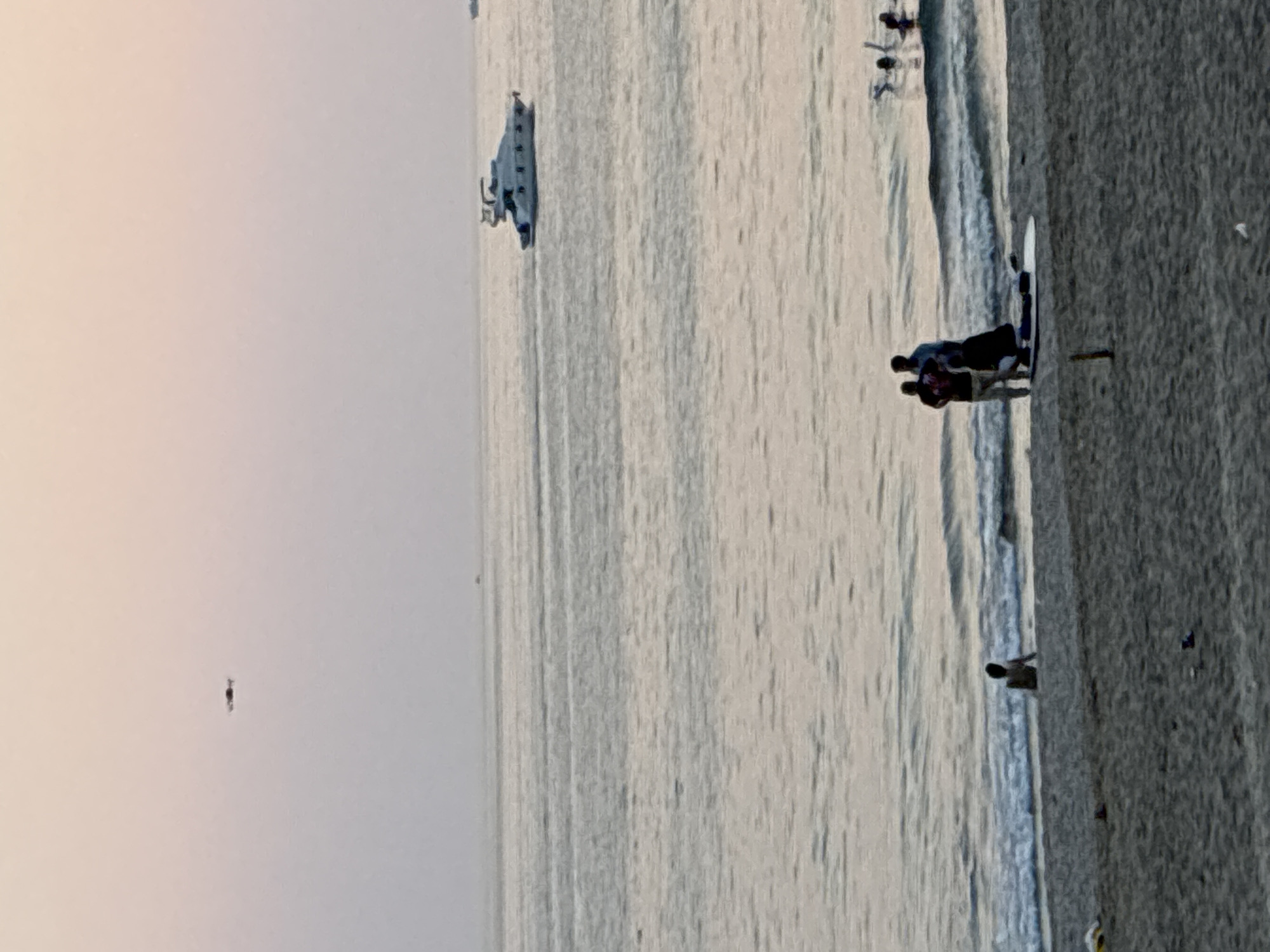 A wide-angle, low-light, aerial shot depicts a serene twilight beach scene. A drone hovers in the soft pink and purple sky. In the distance, a large white yacht navigates the calm sea. Closer to the shore, several figures are present. A lone individual sits at the edge of the water. On the sand, a group of three people are gathered, one of whom appears to be kneeling beside a surfboard. Further out in the water, several more individuals are enjoying themselves. The beach itself is sandy with some scattered debris. This scene is consistent with the coastline of Alanya, Turkey, known for its beautiful beaches and recreational water activities.