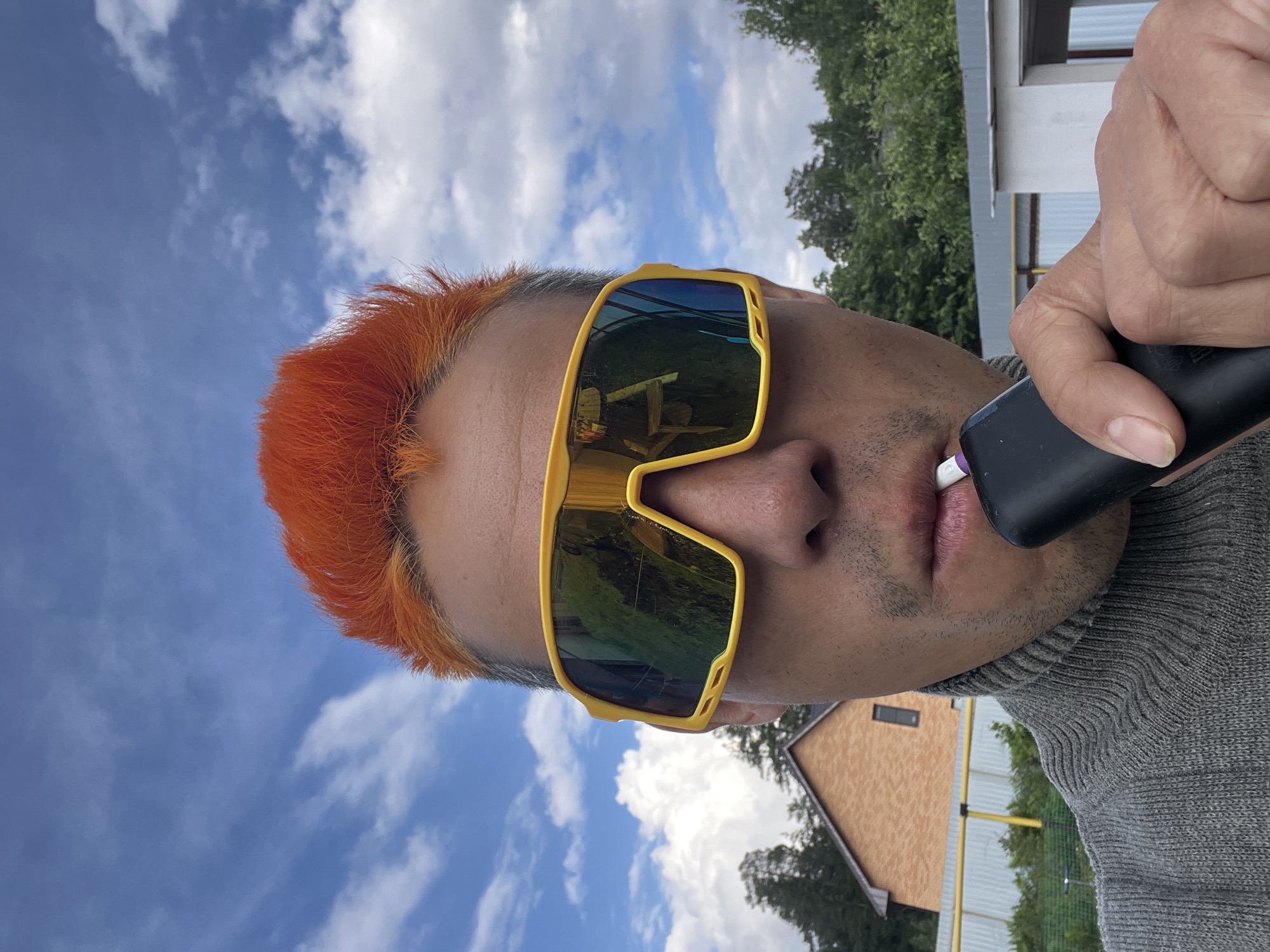 A man with orange hair and yellow-framed sunglasses is smoking an electronic cigarette. The location appears to be outdoors, near buildings and trees. The sky is visible with scattered clouds. There is no specific information that identifies the location as Chaikovsky, Russia.