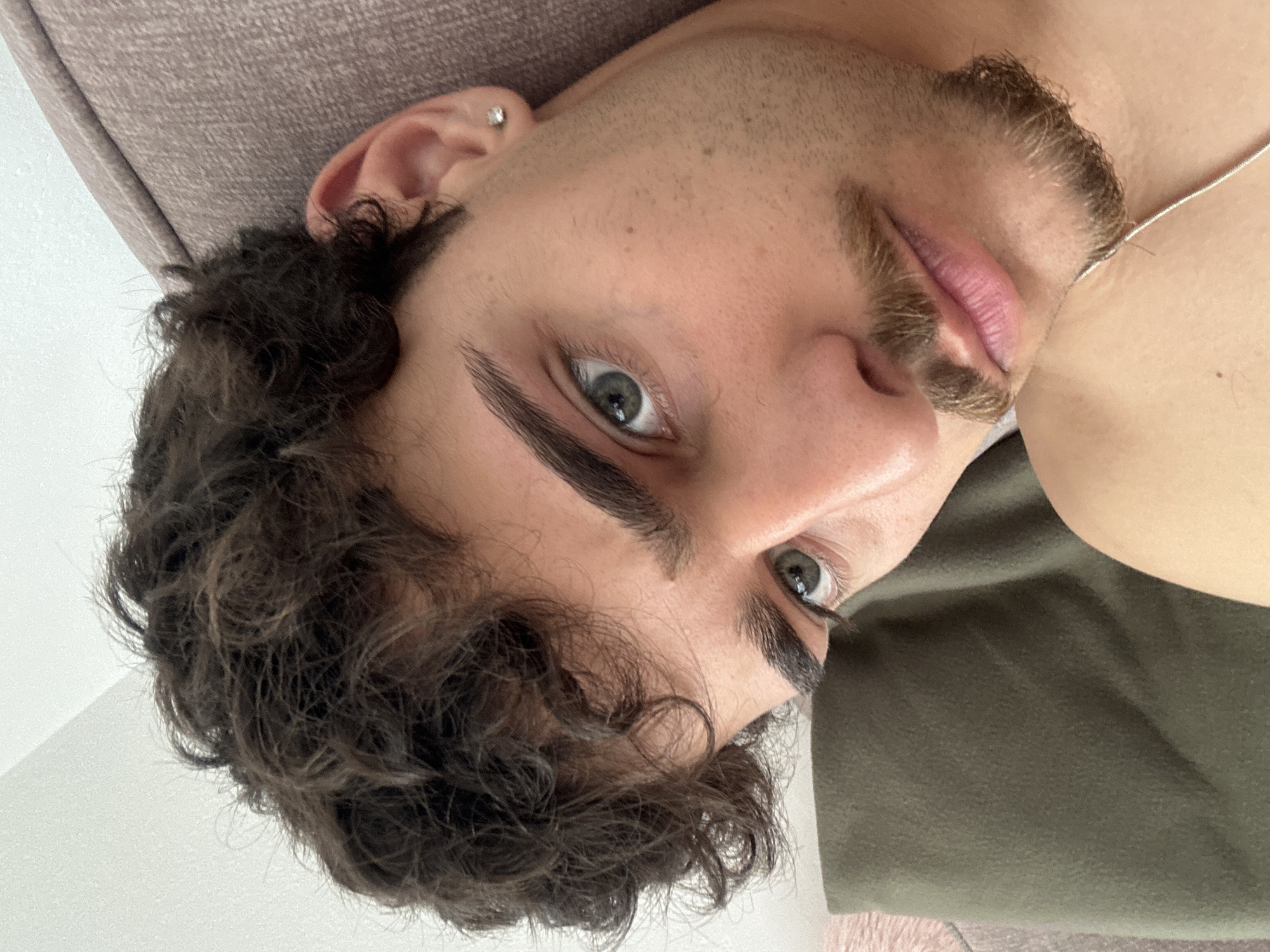 A close-up, slightly angled portrait of a young man with curly dark brown hair and stubble. He has fair skin, well-defined eyebrows, and light gray eyes. A silver earring is visible in his left ear. He appears to be lying down on a gray and olive-colored surface, possibly a couch or bed. Part of a silver chain is visible near his chest. No details suggest Troyan, Bulgaria.