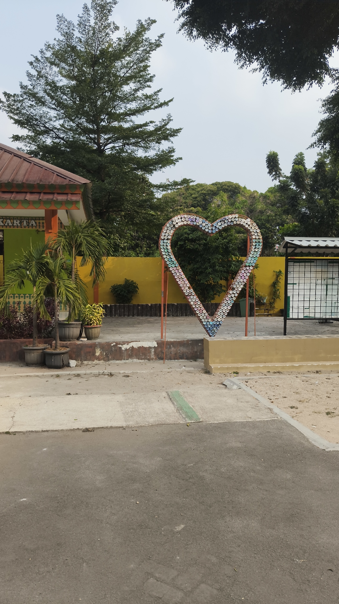 The image shows an outdoor scene in South Jakarta, Indonesia. In the foreground, there is an asphalt road with some debris. Beyond the road, there is a paved area with a concrete walkway that has a green stripe. In the center of the paved area, there is a large heart-shaped sculpture made of plastic bottles. Behind the sculpture, there is a bright yellow wall. To the left of the yellow wall, there is a building with a red roof and green walls, decorated with orange and green trim. Several potted plants, including palm trees, are placed in front of the building. To the right of the heart sculpture, there is a white structure with a grid pattern, possibly a bulletin board or display case. The background is filled with lush green trees and a cloudy sky. The overall impression is that of a public space, possibly a park, school, or community area.