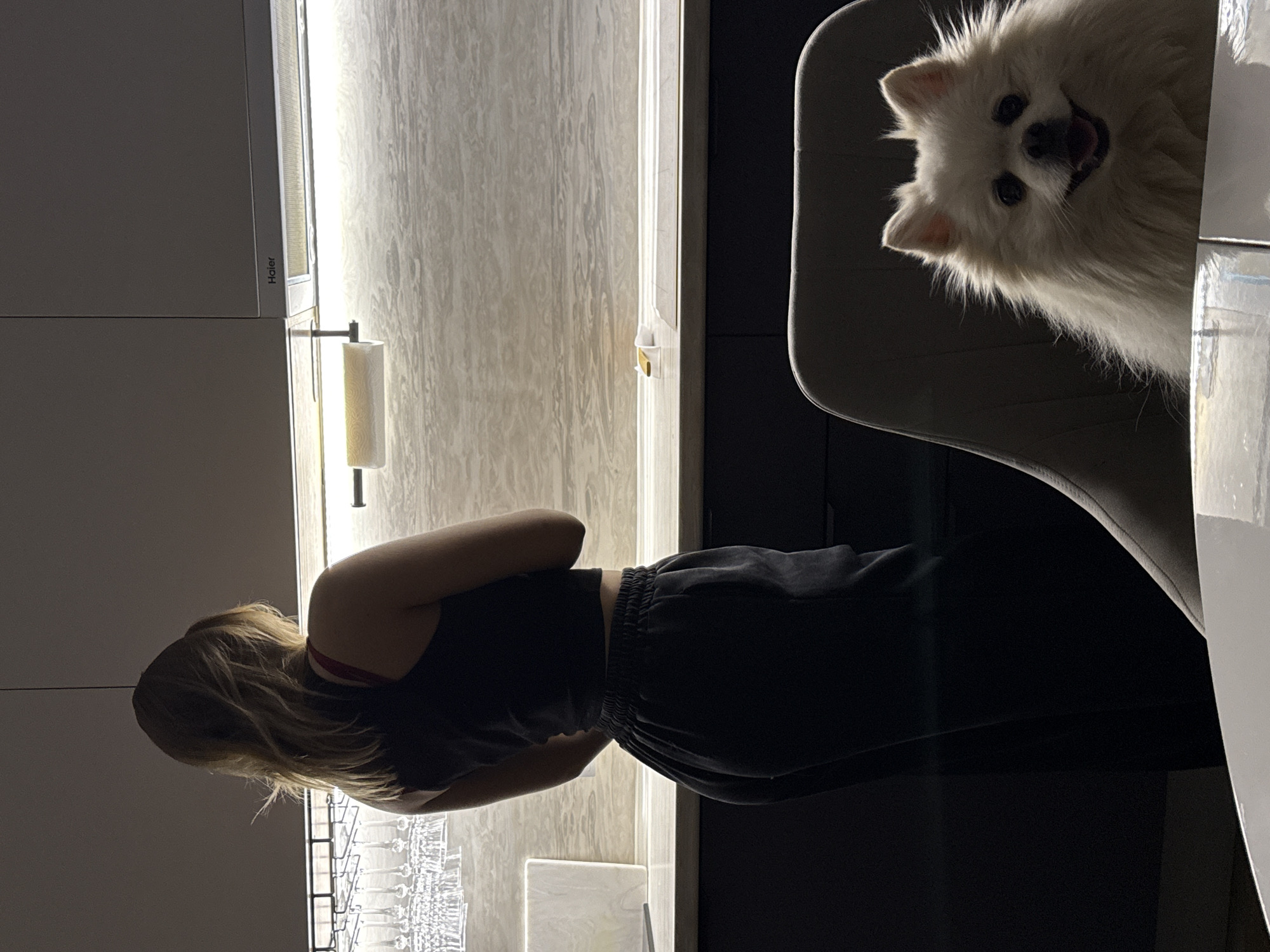 A woman with blonde hair wearing a black tank top and black sweatpants stands indoors near a wall displaying glassware. To her right, a white dog is partially visible, sitting in a gray chair. The location is indoors and appears to be in Лобаново, Russia. The environment seems to be a kitchen or dining area, with modern cabinetry and lighting.