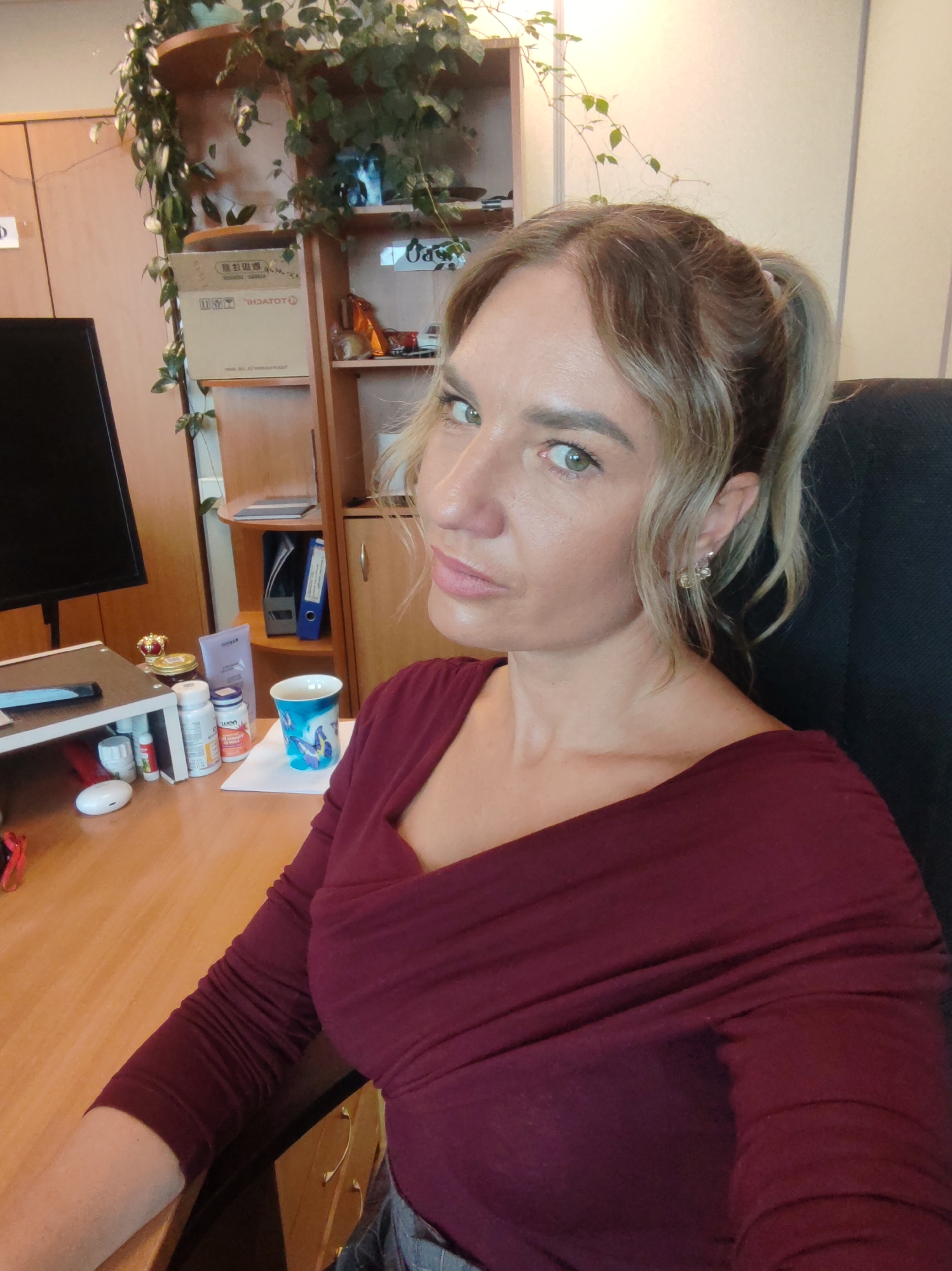A woman is seated at a desk in an office. She is wearing a burgundy long-sleeved top and has her blonde hair pulled back into a ponytail. Her desk has a computer monitor, a mouse, a mug, and several small bottles of pills. Behind her is a wooden bookcase with a plant on top. The location cannot be determined from the image.
