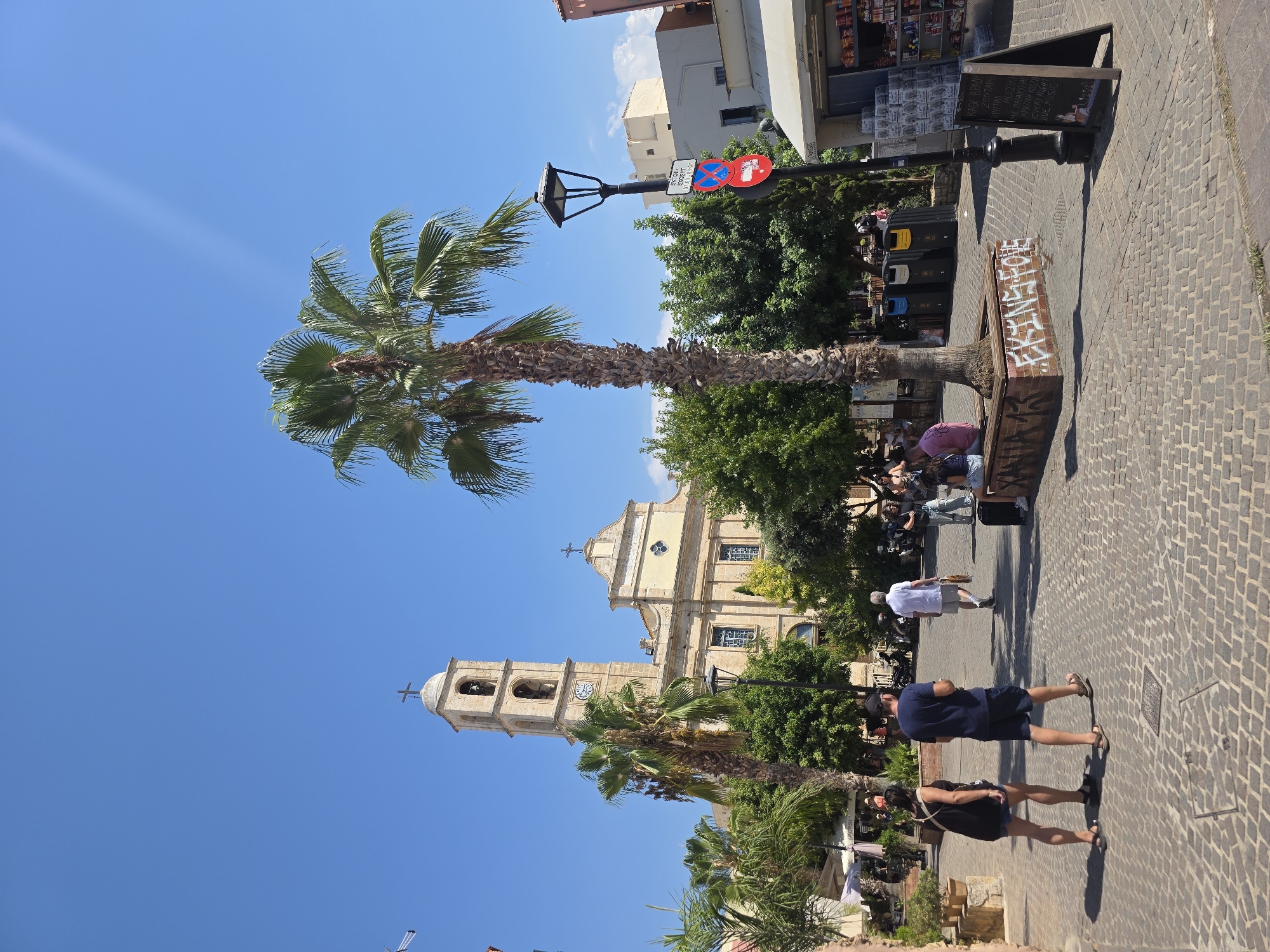 In Chania, Greece, a sun-drenched plaza features a prominent bell tower of the Church of Agios Nikolaos. The square is paved with cobblestones and dotted with palm trees and lush green foliage. A tall, slender palm tree stands near the foreground, its trunk adorned with graffiti that reads "EKINGS LOVE". Several people are present: a couple walks away from the camera in the foreground, dressed in casual summer attire; other individuals are seated on a brick bench near the palm tree. In the background, more people stroll through the square. A streetlamp with signage stands to the right of the palm tree, and a kiosk selling goods is visible. The sky above is a clear, vibrant blue.