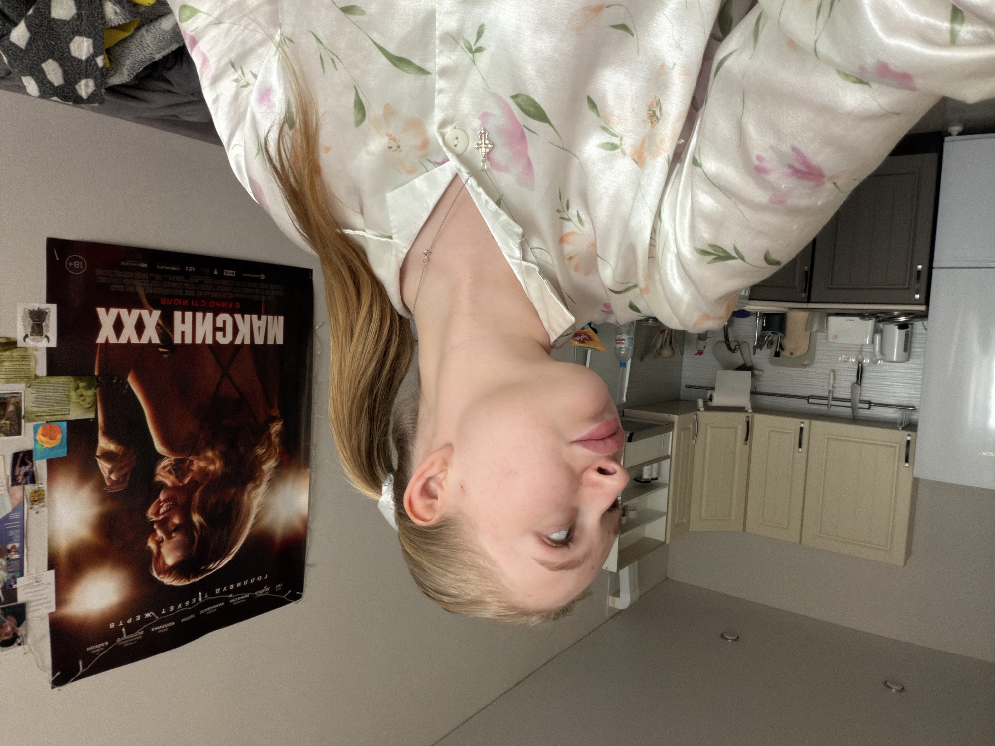The image captures an indoor scene, likely within an apartment or home in Zasechnoye, Russia. The primary subject is a young woman, partially visible, centered in the frame. She has fair skin and light blonde hair pulled back into a ponytail. She is wearing a light-colored pajama top, possibly satin, adorned with a delicate floral pattern in shades of pink and green. A silver cross necklace is visible around her neck, with another small cross charm pinned to her top's collar. Her gaze is directed slightly upwards and to her right, and her expression is neutral.
The setting appears to be a combined living space. To the left of the woman, a wall features a movie poster for "МАКСИН XXX" (MAXINE XXX) with an "18+" rating and the text "В КИНО С 11 ИЮЛЯ" (IN CINEMA FROM JULY 11). Below the movie title, the phrase "ГОЛЛИВУД ТРЕБУЕТ ЖЕРТВ" (HOLLYWOOD DEMANDS SACRIFICES) is visible. Several smaller photos or notes are pinned to the wall next to the poster. Above this, a pile of clothes or textiles, including one with a black and white polka dot pattern, is resting on a surface.
To the right and in the background, a kitchen area is visible. It features light-colored base and wall cabinets with dark handles, a sink area with a faucet, and various kitchen items on the countertop, including a water bottle and utensils. A white refrigerator or large appliance stands at the far right. The floor is a light, neutral color. The lighting suggests an indoor daytime or evening setting. The overall atmosphere is personal and domestic.