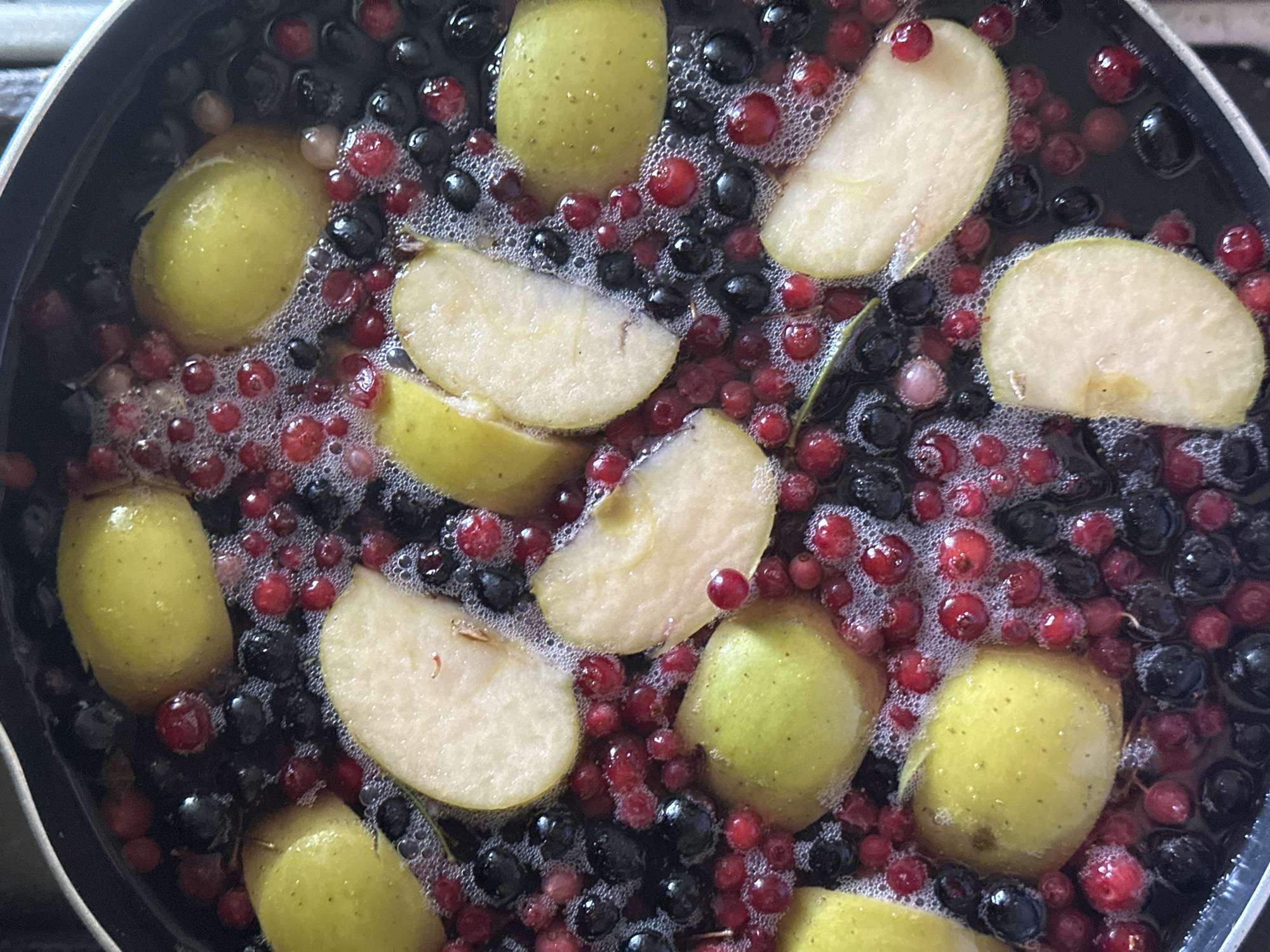 The image shows a metal pot filled with a mixture of red and black berries, along with slices and whole pieces of light green apples, all submerged in a liquid that is bubbling slightly. The ingredients appear to be in the process of being cooked or prepared, possibly for a jam, compote, or similar dish. The setting is indoors, likely in a kitchen or cooking area. There is no indication of the location being Nizhny Novgorod, Russia. No people, subjects, objects, actions, interactions, and the environment are visible.