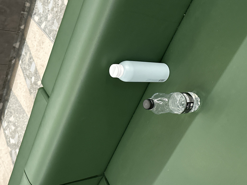 Two bottles rest on a green, textured surface. One is a pale blue, opaque bottle with a white cap. Next to it, a clear plastic water bottle with a black cap appears partially crushed. The bottles are oriented horizontally on the green surface, which appears to be upholstered. The left side of the frame reveals a glimpse of a patterned, tiled floor.