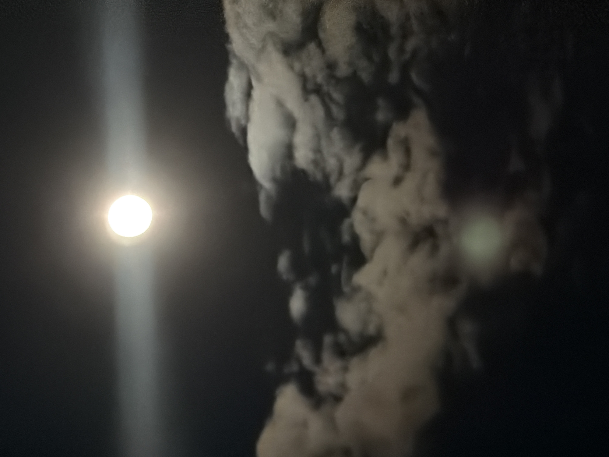 The image presents a dark night scene. On the left side of the frame, a bright circular object, presumably the moon, emits a strong beam of light extending vertically. To the right, there are cloudy forms that appear illuminated in parts, contrasting with the surrounding darkness. The location (Chaikovsky, Russia) and any further context beyond what is visible in the image are not discernable.