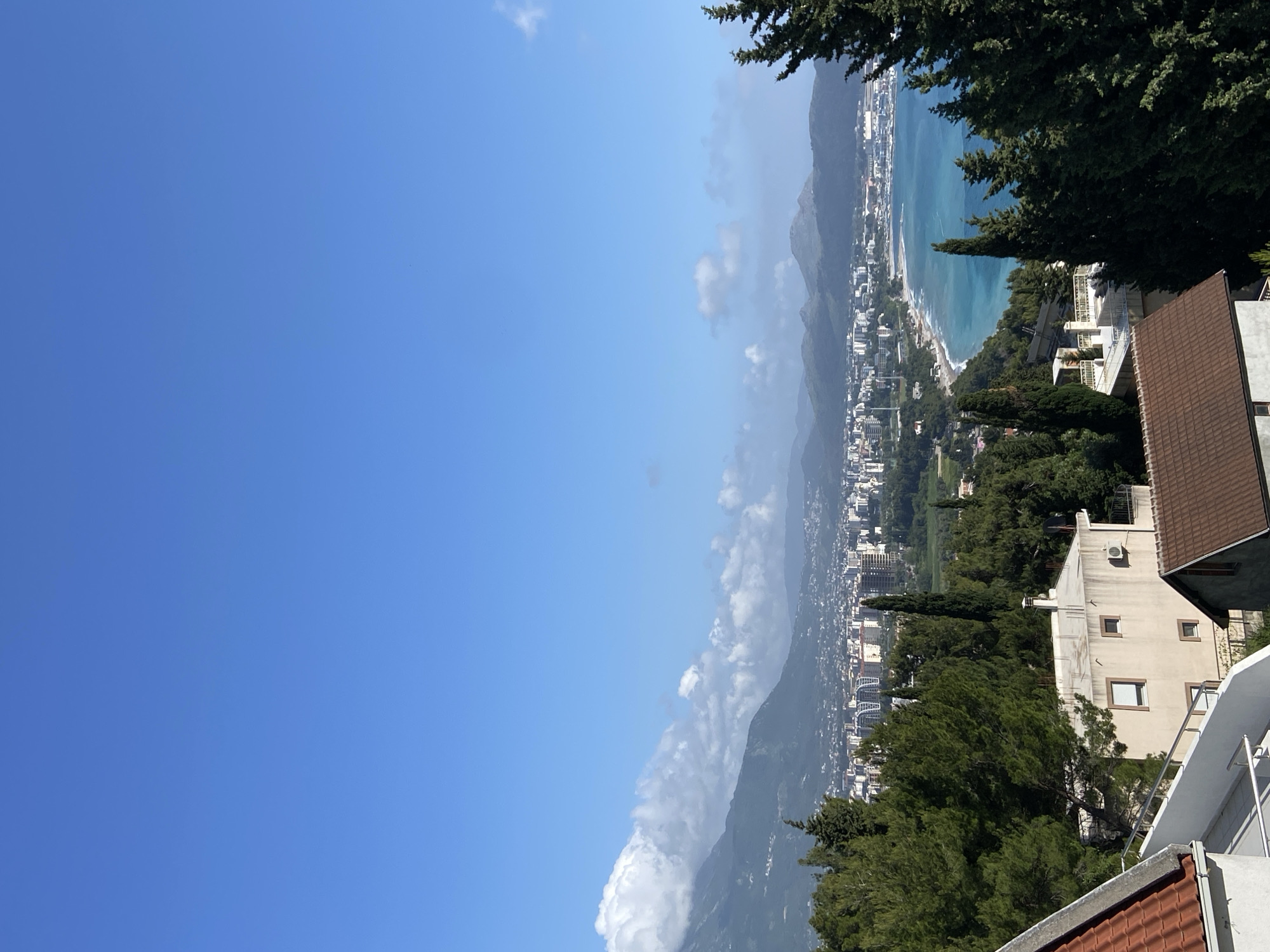 This is an aerial view of Šušanj, Montenegro. The Adriatic Sea stretches along the coastline. In the foreground, there are trees and residential buildings. In the background, a mountain range is partially obscured by clouds. The sky is clear blue with some scattered white clouds. The photograph has been taken in the daytime. The image is rotated counterclockwise.