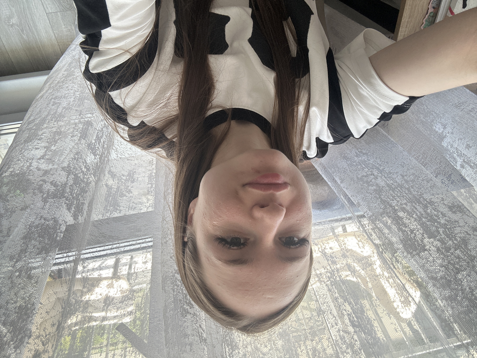 A young woman is depicted in a close-up, head-and-shoulders selfie shot, with her head positioned towards the bottom of the frame and her body extending upwards (when viewed upright, not rotated). She looks directly at the camera with a neutral expression. Her long, dark brown hair falls around her face and over her shoulders. She is wearing a short-sleeved top with a distinct black and white pattern, resembling cow print or abstract shapes. Her eyelashes are long, and her lips are slightly parted.
The setting appears to be indoors, likely a room with a large window. Behind her, sheer, light-colored curtains with an intricate, textured pattern are drawn, filtering the bright light coming from outside. Through the curtains, hints of an outdoor environment with trees or greenery are visible, indicating it is daytime. A wooden or laminate floor is visible on the upper left side of the frame, and a brown, possibly wooden, furniture piece is partially seen on the upper right. There is no visible text in the image.