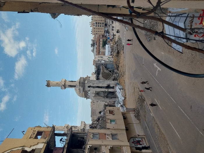 The image displays a street scene in Khan Yunis, Palestinian Territory, under a blue sky with scattered white clouds, characterized by widespread urban destruction. The prominent structure in the mid-ground is the Al-Farouq Mosque, identified by its distinctive minaret and dome, which shows severe damage including collapsed sections and surrounding debris. To the left, a multi-story building is significantly damaged, revealing exposed interiors, rebar, and rubble. The asphalt road features white lane markings, including a double solid line and a right-turn arrow. Four individuals, appearing to be children, are actively playing football in the street; one is wearing a red shirt. Further down the street, approximately eight pedestrians are visible walking. The immediate foreground on the right side of the frame shows a section of a damaged building's wall with exposed rebar and tangled wires. Piles of construction debris, concrete, and sand are scattered along the roadside and around the damaged structures.