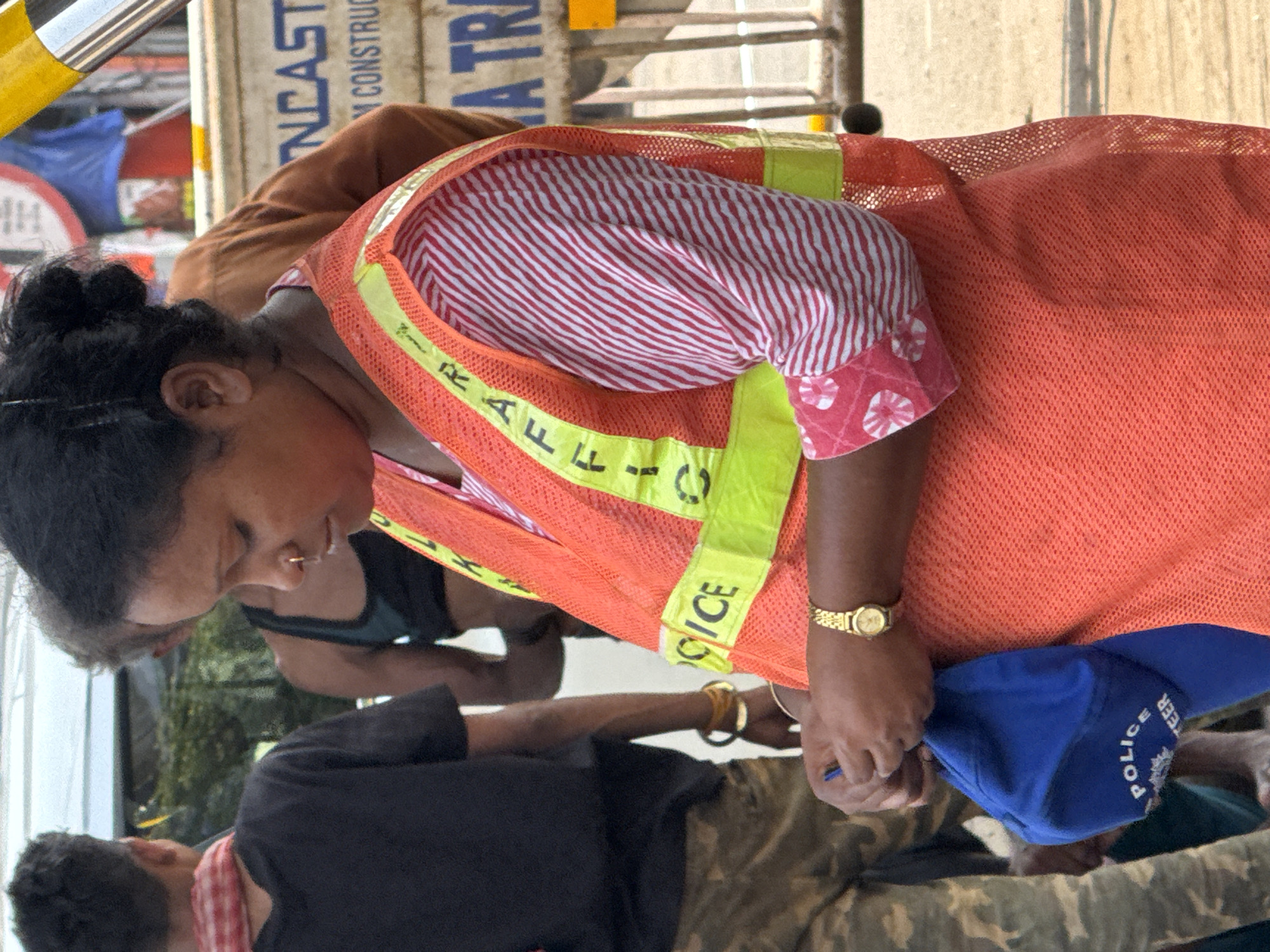 In Kolkata, India, the image depicts a woman with dark hair wearing a coral-colored mesh safety vest with "TRAFFIC POLICE VOLUNTEER" printed on it. Underneath the vest, she wears a white shirt with red stripes and pink polka-dot cuffs. She is holding a blue "POLICE VOLUNTEER" baseball cap. She has a nose piercing and a gold wristwatch. Several people are visible behind and around her, including a man in a black t-shirt and camouflage pants. The background includes construction-related materials and signs, indicating an urban environment.