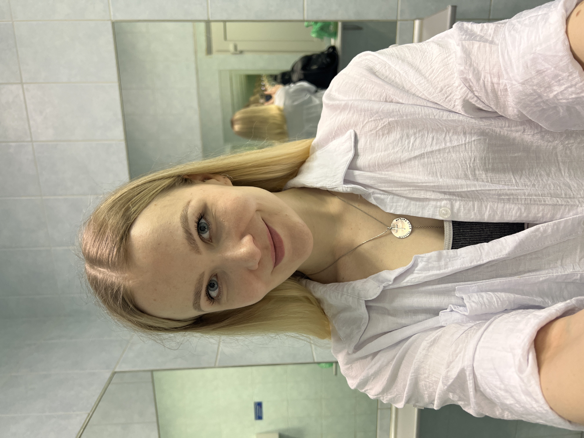 The image shows a young woman with fair skin, blonde hair, and blue eyes, taking a selfie in a bathroom. She is wearing a white, collared, button-down shirt and a silver necklace with a circular pendant. In the background, a mirror reflects the bathroom interior, including tiled walls and other indistinct bathroom fixtures. A glimpse of another person is visible in the mirror's reflection. The location is identified as Khimki, Russia.