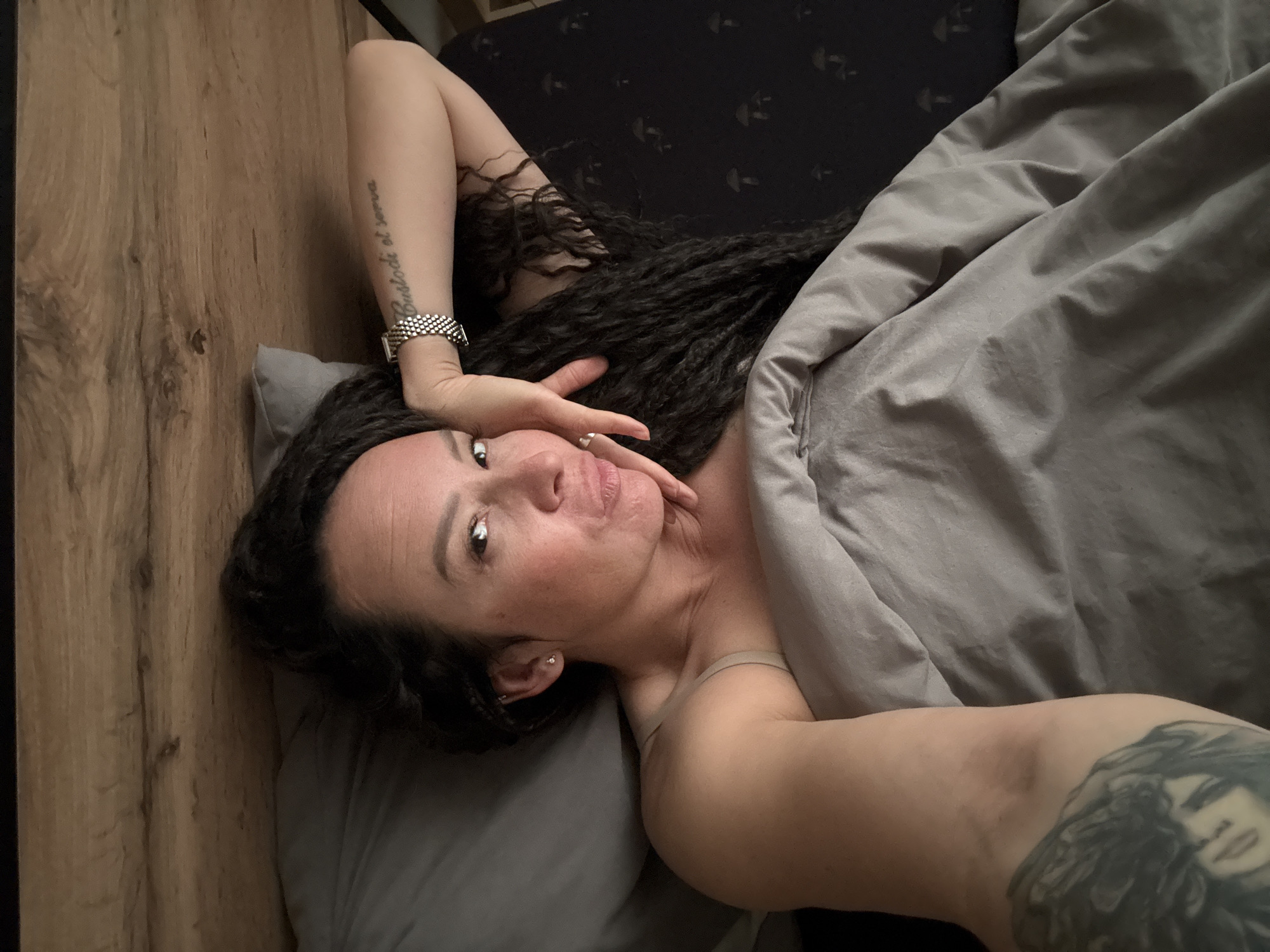 A woman lies in bed, looking directly into the camera with a slight pout. She has long, dark, textured hair. Her right arm is bent, with her hand resting under her chin. A metallic link-style watch and a tattoo reading "Custodi et serva" are visible on her right forearm. A ring is present on the ring finger of her right hand. Her left upper arm displays a large, dark tattoo of a stylized face. She is partially covered by a light grey duvet, and a light-colored strap is visible on her left shoulder. A grey pillow supports her head. To her right, a dark blue or black pillow with light-colored mushroom patterns is visible. The bed features a light brown, wooden headboard with a distinct wood grain. The indoor setting, consistent with a bedroom, contains no visual information to indicate its specific geographical location, nor does it provide any evidence to place it in the city of Chaikovsky, Russia.
