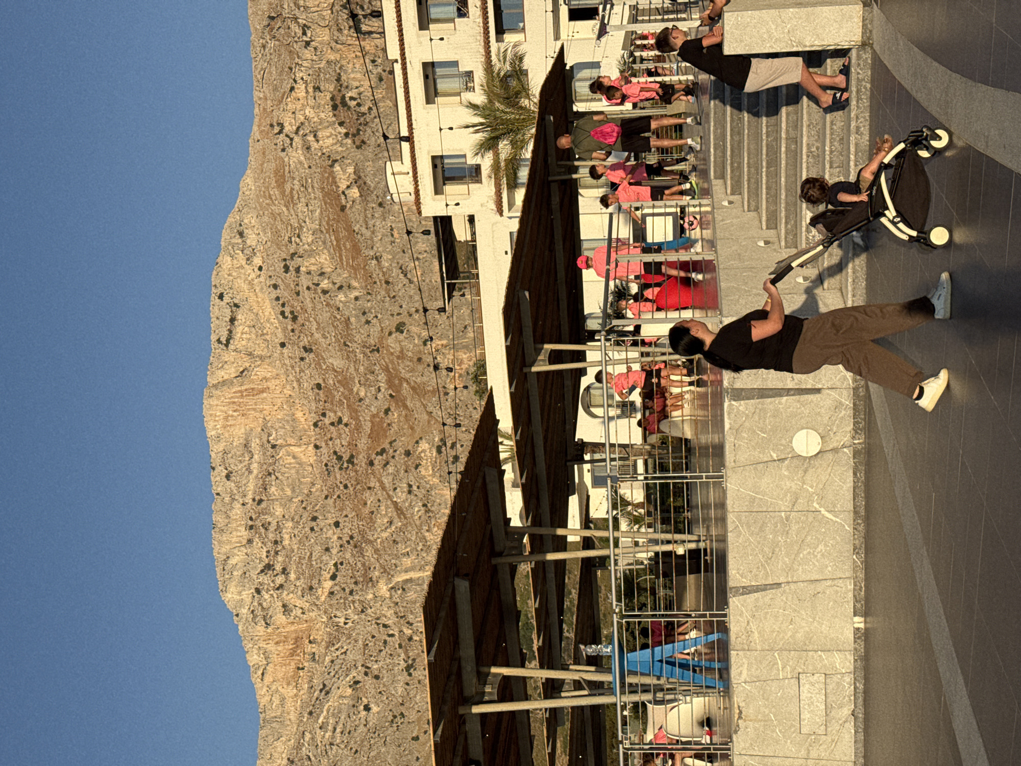 In Capaci, Italy, on a sunny day, a woman pushes a stroller containing a child along a paved walkway. Behind them, a group of children and adults, many wearing pink attire, are gathered on a raised area with a building in the background. A large, rocky mountain dominates the skyline. The scene appears to be a public space, possibly a promenade or plaza, adjacent to a coastal resort.