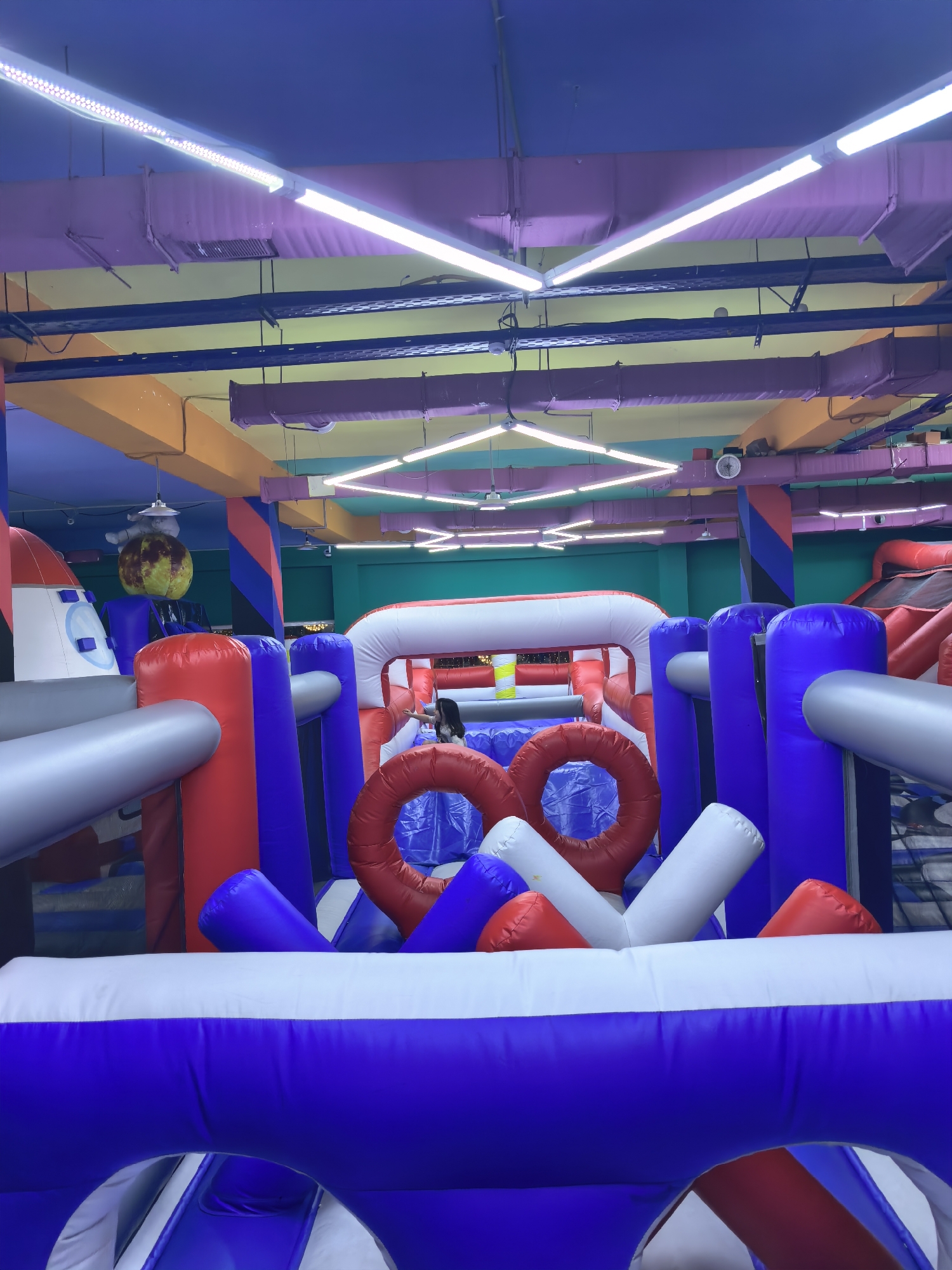 The image depicts an indoor inflatable playground, possibly a family entertainment center or a children's play area. The environment is brightly lit by fluorescent lights, and the ceiling is painted in vibrant colors of purple, yellow, and orange, with exposed ventilation systems. Dominating the foreground and mid-ground is a large, multi-colored inflatable structure composed of red, white, and blue sections. This structure features various elements such as inflatable columns, arches, tunnels, and what appear to be rings or obstacles. In the background, more inflatable structures are visible, including a red inflatable slide or climbing area. To the left, part of a rocket-shaped inflatable is visible, along with a spherical inflatable resembling a planet. A young girl is partially visible on the right side of the image, interacting with the inflatable structures. The overall impression is one of a playful and energetic indoor recreational space. The image does not provide any explicit information or visual cues to suggest the location is Fergana City, Uzbekistan.