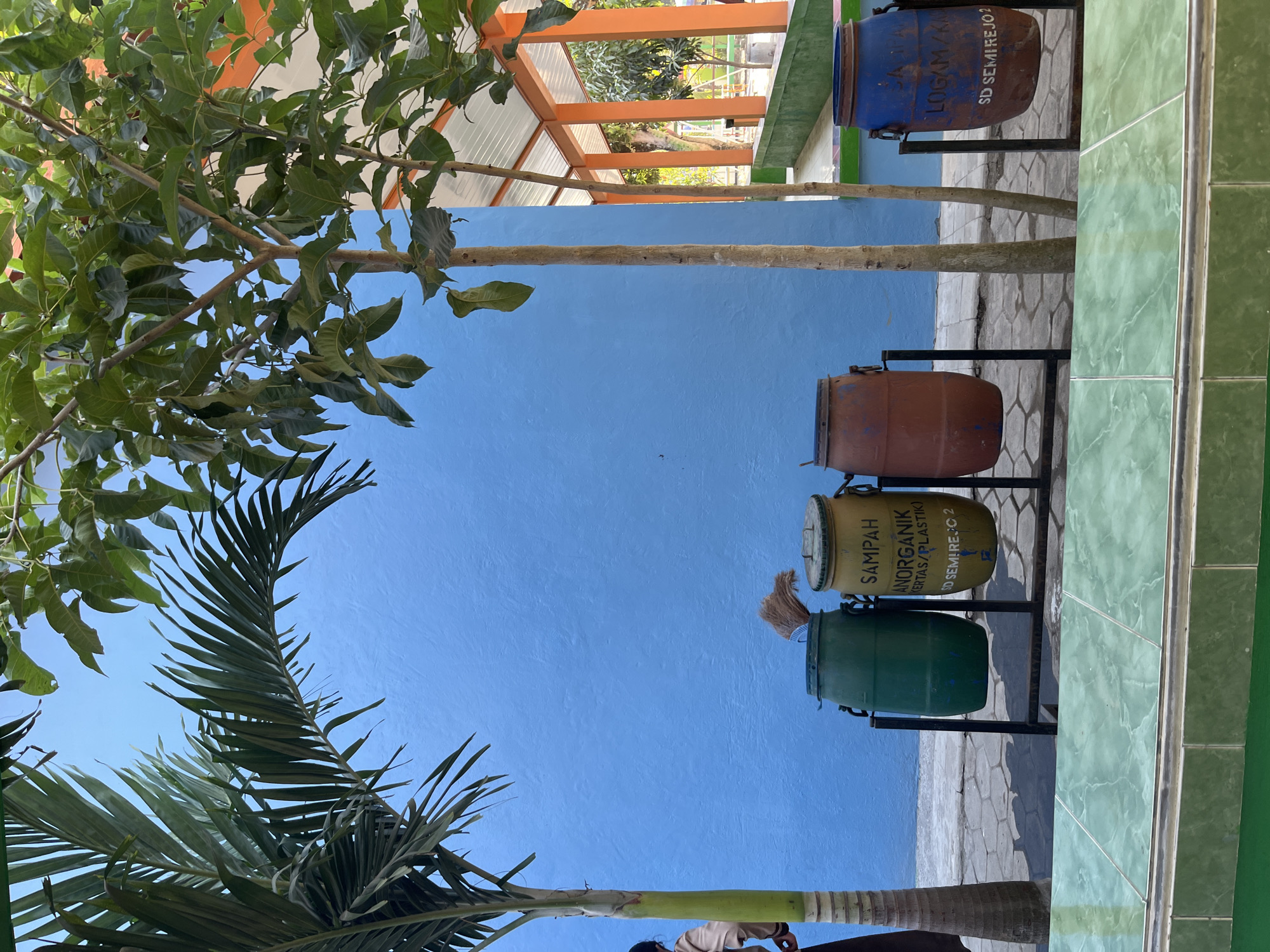 In Gembong, Indonesia, a row of four large, repurposed plastic barrels are positioned outdoors against a vibrant blue wall. From left to right, the barrels are green, yellow, reddish-brown, and blue. Each barrel is mounted on a black metal stand. The yellow barrel has text printed on it, reading "SAMPAH ANORGANIK (KERTAS/PLASTIK) SD SEMIREJO 2". The blue barrel has similar text, "SAMPAH LOGAM/K.... SD SEMIREJO 2". A broom with a bundle of straw is leaning against the inside of the green barrel. To the left of the barrels, a person in a brown jacket and dark pants stands near the base of a tall palm tree. The palm tree's fronds extend towards the top of the image. Two slender trees with light brown bark and green leaves stand between the barrels, with the reddish-brown and blue barrels partially obscured by them. In the background, a covered walkway with orange trim and a white roof extends to the right. The ground is paved with hexagonal light gray stones. A low green tiled wall runs across the bottom foreground of the image.