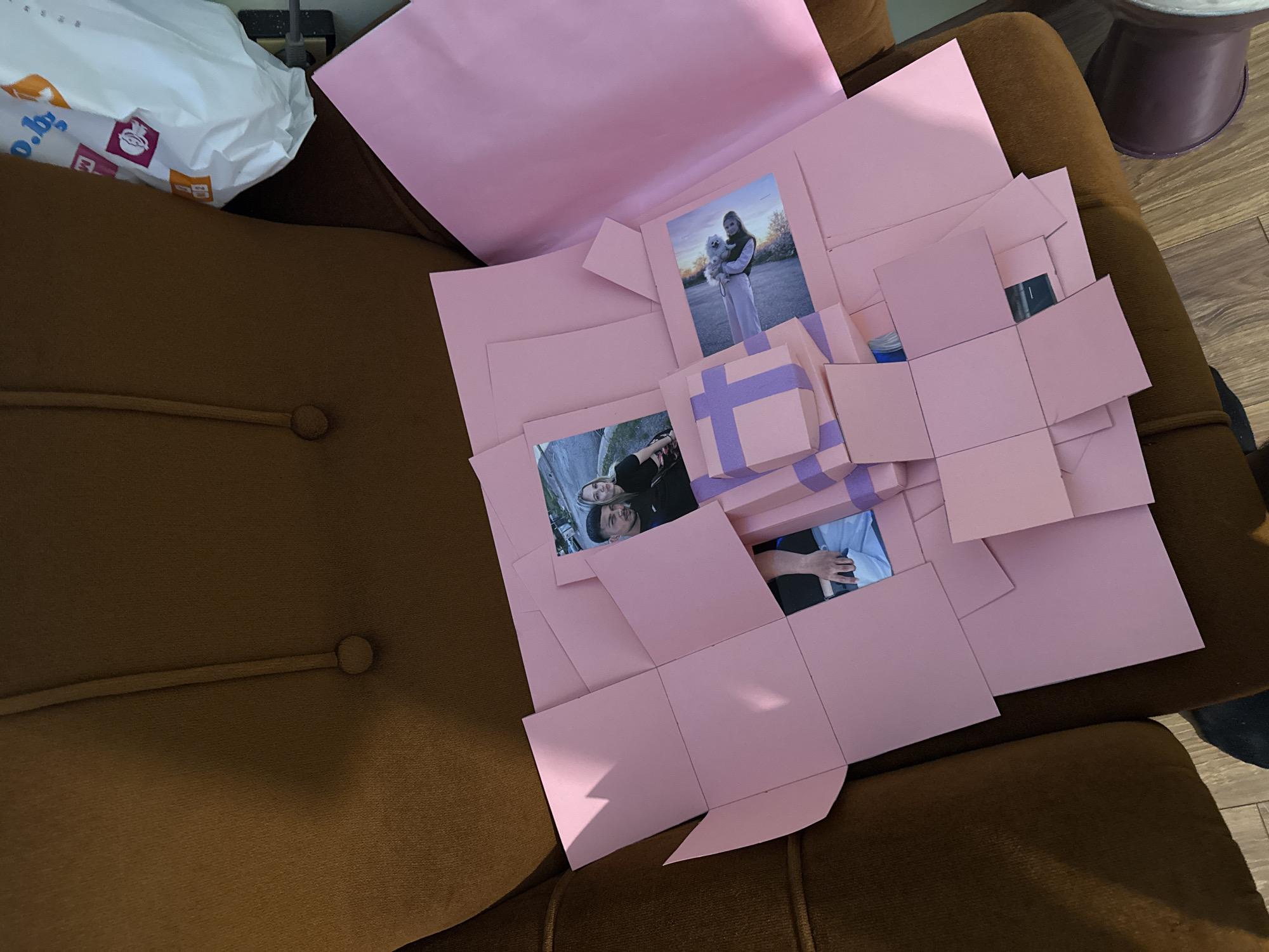 An open, light pink explosion box rests on a brown upholstered armchair or sofa. The multi-layered craft box, made from cardstock, has unfolded panels revealing its contents. In the center, several smaller pink gift boxes decorated with purple ribbon designs are stacked.
Various personal photographs are affixed to the internal panels of the explosion box. One prominent photo on the upper right depicts a woman with long blonde hair holding a small white fluffy dog, likely a Pomeranian, outdoors during what appears to be golden hour with soft, warm lighting. Another photo on the middle left shows a smiling couple, a blonde woman and a bearded man, embracing, with a blurred outdoor background. A third, partially visible photo at the bottom right shows a person's hand or arm wearing a light blue sleeve.
The setting appears to be indoors, likely a casual living space in Yana, Bulgaria. In the upper left background, part of a white plastic bag is visible with fragmented text, including "o.by" and a purple square logo. A dark object, possibly an electronic plug, and a grey cord are also seen behind the bag. On the upper right, a section of a dark-colored stool or table leg is visible, resting on a wooden floor. The explosion box, often used for gifting, suggests a celebration or commemoration of personal memories, conveyed through the happy scenes in the photographs.