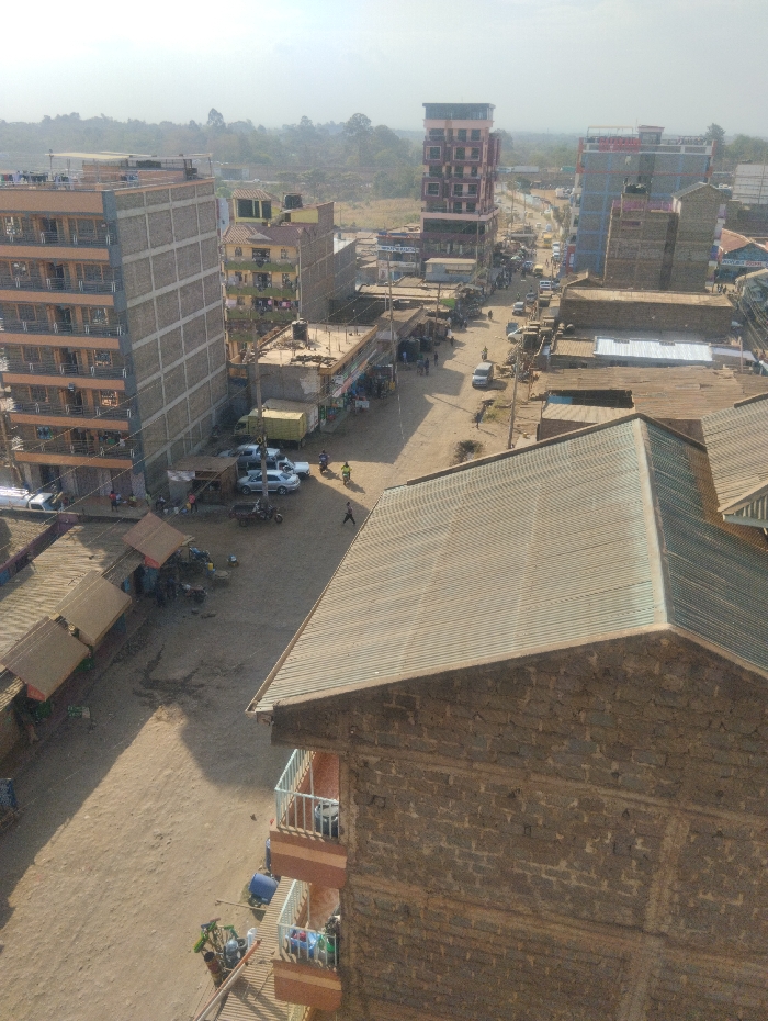 This image presents an elevated view of a bustling urban environment within Witeithie ward, Kenya. The scene is dominated by a central, unpaved, light brown dirt road that extends into the hazy distance, flanked by a dense arrangement of buildings.
In the immediate foreground on the right, the corrugated iron roof and part of the stone-faced exterior of a multi-story building are visible, featuring balconies with white railings containing blue barrels and laundry. To the left, a parallel dirt path hosts several small market stalls with brown canvas awnings, alongside parked motorcycles and a few white cars.
The midground features the main dirt road, on which several vehicles including light-colored cars, motorcycles, and a yellow truck are present. Numerous pedestrians are observed walking along the road and adjacent paths. The road is bordered by a variety of structures: multi-story buildings ranging from three to seven floors, some with exposed concrete frameworks and orange-painted facades, indicating ongoing development or typical construction. Many rooftops feature corrugated iron and visible water storage tanks.
Further along the main thoroughfare, a prominent tall, narrow building with a reddish-brown exterior stands out, and another multi-story structure with a light blue and grey facade is visible. The background shows the urban density gradually thinning, transitioning into a landscape with sparse trees and scrubland under a bright, hazy sky. The overall environment depicts a dynamic urban landscape with active street life and a mix of residential and commercial structures.