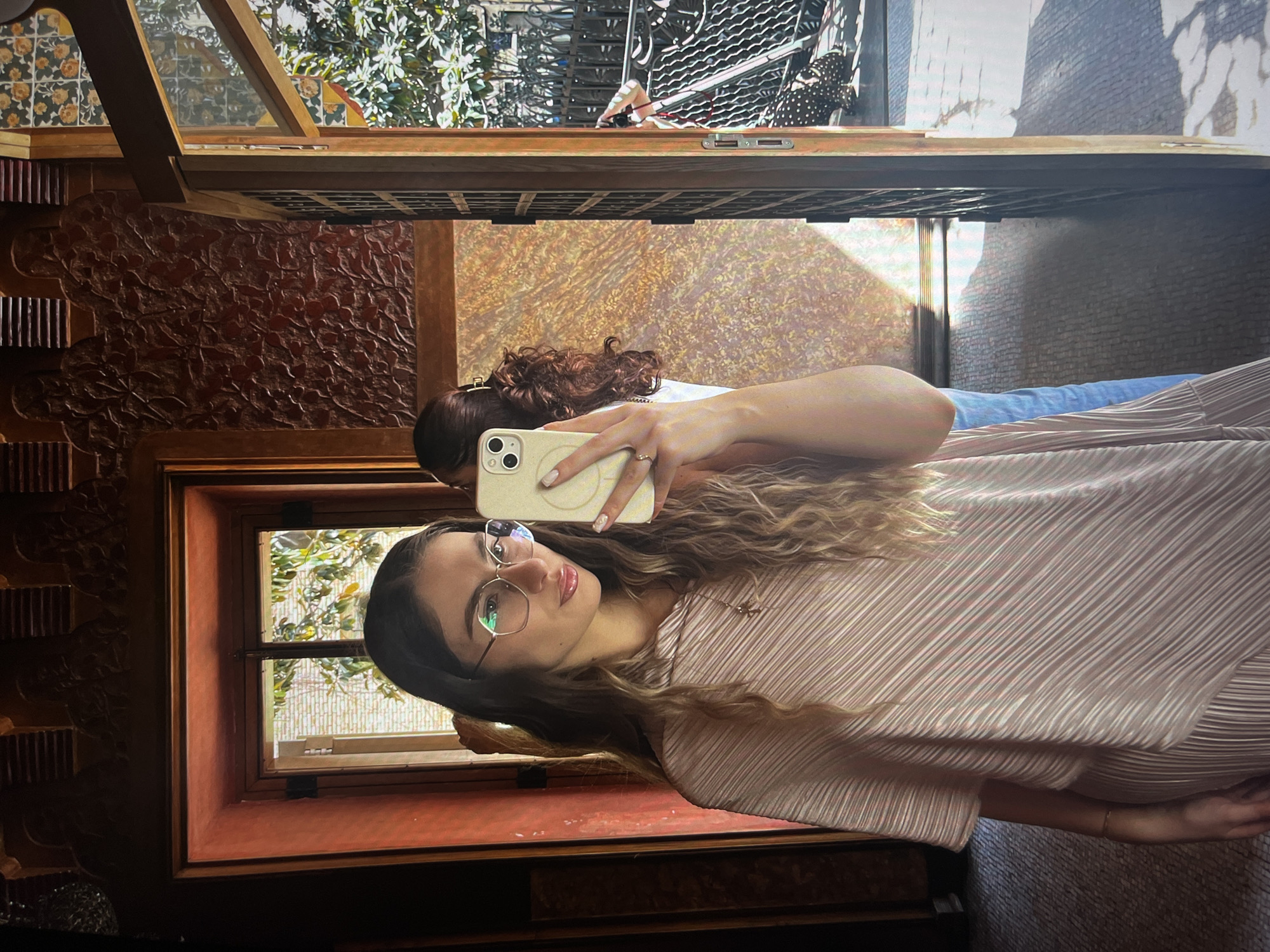 Two women are taking a selfie inside a decorative building. One woman is holding a cell phone and is facing the camera wearing glasses and a pink pleated dress. Another woman with curly hair is partially visible behind her and also holding a phone. There is a window with a view of trees behind the primary woman. The location is the city of Unknown, Unknown.