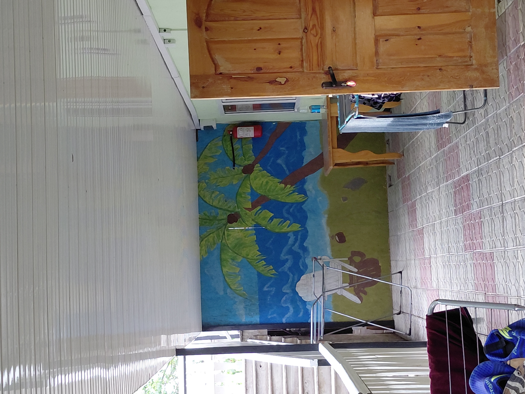 The image depicts an outdoor covered patio area. The ceiling is made of white, horizontal panels. A mural on the back wall shows a tropical scene with palm trees, a blue ocean, and a green grassy area. In front of the mural, there is a white Adirondack-style chair and a portable drying rack with laundry. To the right, a wooden door with vertical panels and a dark handle is partially open. A red fire extinguisher is mounted on the wall near the door. The floor is tiled with a pattern of gray and pink squares. On the left side of the image, a white railing and a set of outdoor stairs are visible. Another drying rack is present in the foreground, holding a dark red piece of fabric and a pair of blue sandals. The location is identified as Ақши, Kazakhstan.