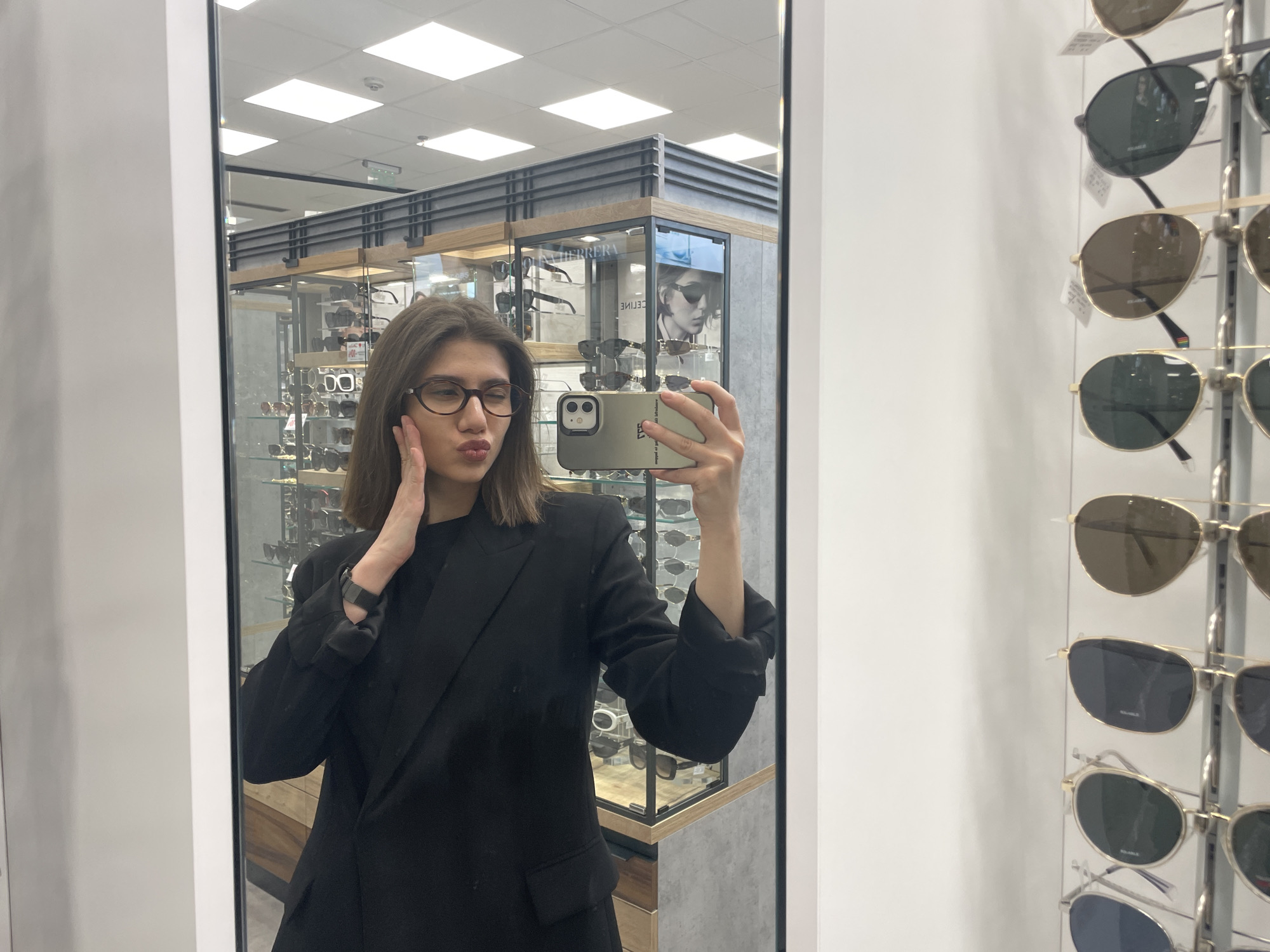 A woman is taking a selfie in front of a mirror inside an eyewear store in Sofia, Bulgaria. She is wearing a black blazer and glasses. She is holding a smartphone in her right hand and touching her face with her left hand, puckering her lips in a kiss. Behind her is a display case filled with eyeglasses and sunglasses. To the right of the mirror is a metal rack holding sunglasses. The lighting is bright and the scene is clean.