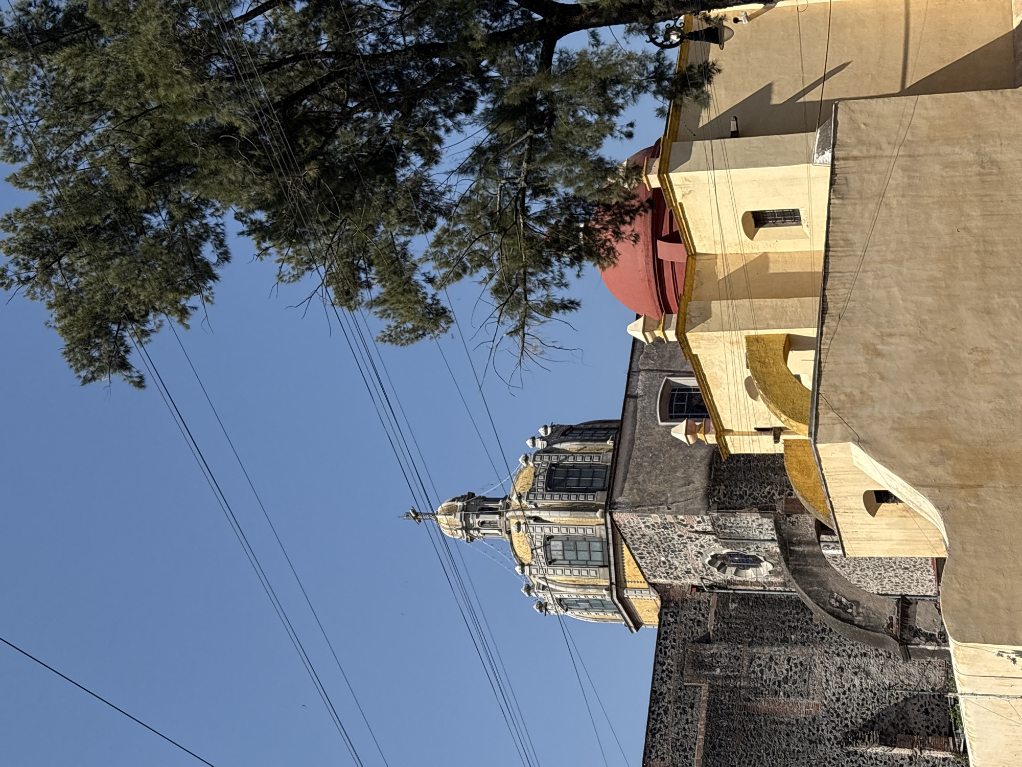 A ground-level, eye-level shot of the Templo de Santa Teresa la Antigua in Mexico City, Mexico, captured on a clear, sunny day. The building, constructed in stages from the 17th to 19th centuries, features stone masonry, plaster, yellow and red paint, and curved architectural details, including multiple domes of varying sizes. Overhead, a tree’s branches stretch across the frame against a clear blue sky. Visible electrical lines add to the scene's complexity. The building's exterior is in a state of preservation, with some wear and tear visible.