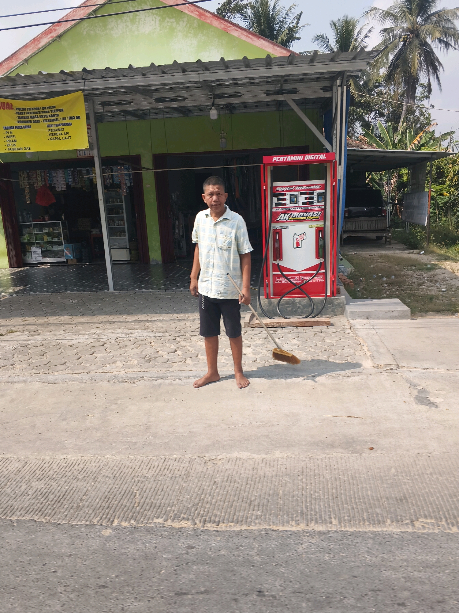 In Jati Indah, Indonesia, a man stands barefoot on a concrete sidewalk, holding a broom. He is dressed in a light-colored checkered short-sleeved shirt and dark shorts. Behind him, a small building with a green facade features a yellow banner listing various services. To his right, a red and white "PERTAMINI DIGITAL" gas pump stands prominently. The environment appears to be a roadside stall or small shop, with vegetation and palm trees visible in the background.