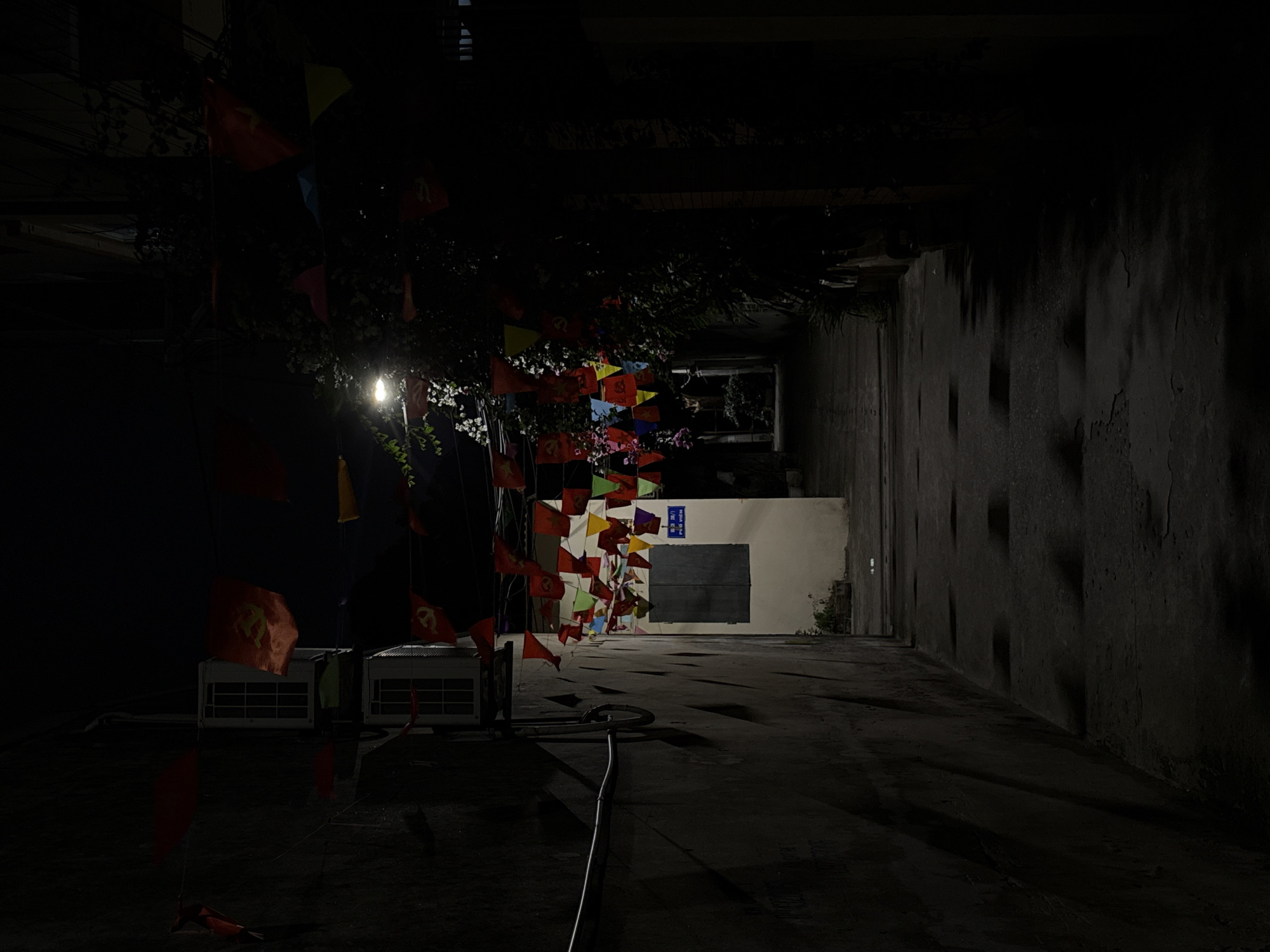 The image shows a dimly lit alley in Hà Nội, Vietnam, at night. Strings of colorful triangular flags are strung across the alley, partially obscuring the view and casting shadows. In the foreground, two air conditioning units sit on the ground. A power cord snakes across the alley floor. The alley walls are dark and appear to be made of weathered concrete. In the background, a white wall with a dark square is visible, with a window above it. The scene appears deserted.