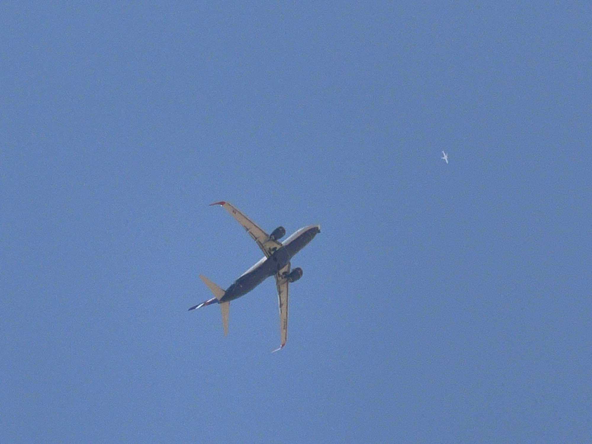 An aerial view captures two airplanes against a clear blue sky. The larger airplane is prominently positioned in the upper left quadrant, viewed from below and slightly from the side, with its wings extended and engines visible. Its tail features a distinctive red, white, and blue design. A second, much smaller airplane is visible in the lower right quadrant, appearing as a tiny white silhouette against the vast blue expanse. The scene is devoid of any identifiable landmarks or contextual information that would suggest its location in Sharm El Sheikh, Egypt.