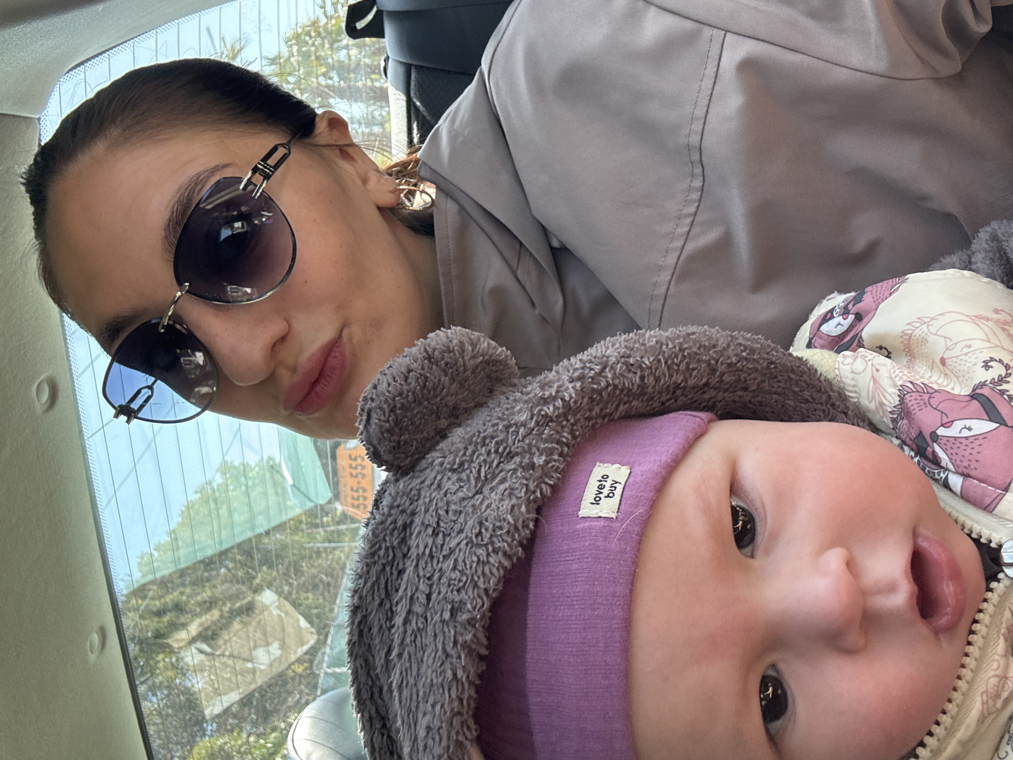 A woman in sunglasses and a grey jacket is taking a selfie with a baby wearing a purple hat and a grey hooded jacket. They appear to be in a car, as evidenced by the rear window and seat. Outside the car window, there are trees and a building, and a sign with "555-555" is visible. The location is Sochi, Russia.