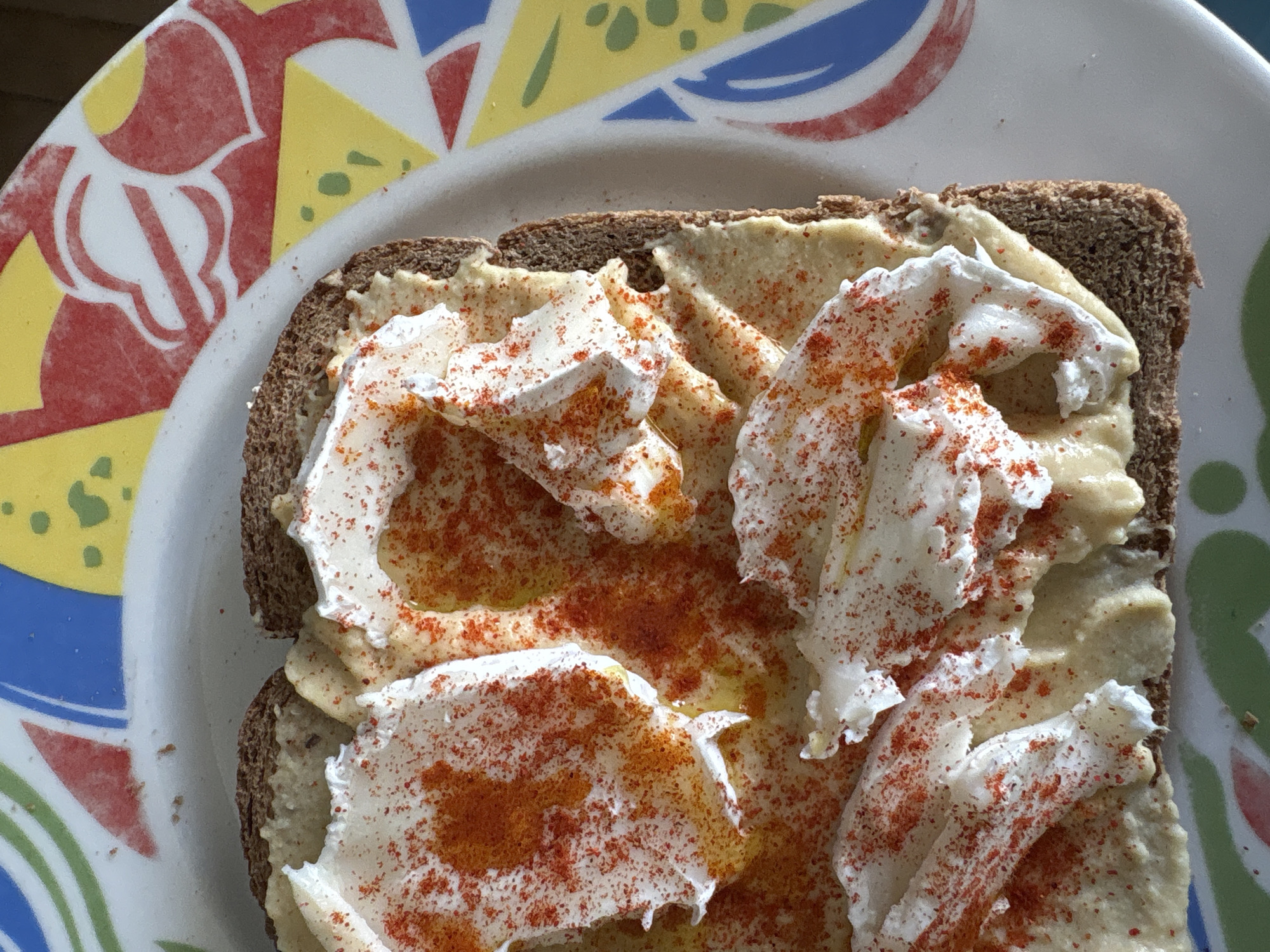 A close-up, high-angle shot depicts a piece of dark brown bread topped with hummus and what appears to be ricotta cheese or a similar creamy topping, sprinkled with paprika and drizzled with oil. The bread and toppings are situated on a plate with a multicolored pattern of red, yellow, blue, and green. The location is Urbanización Encinares De Alcolea, Spain.
