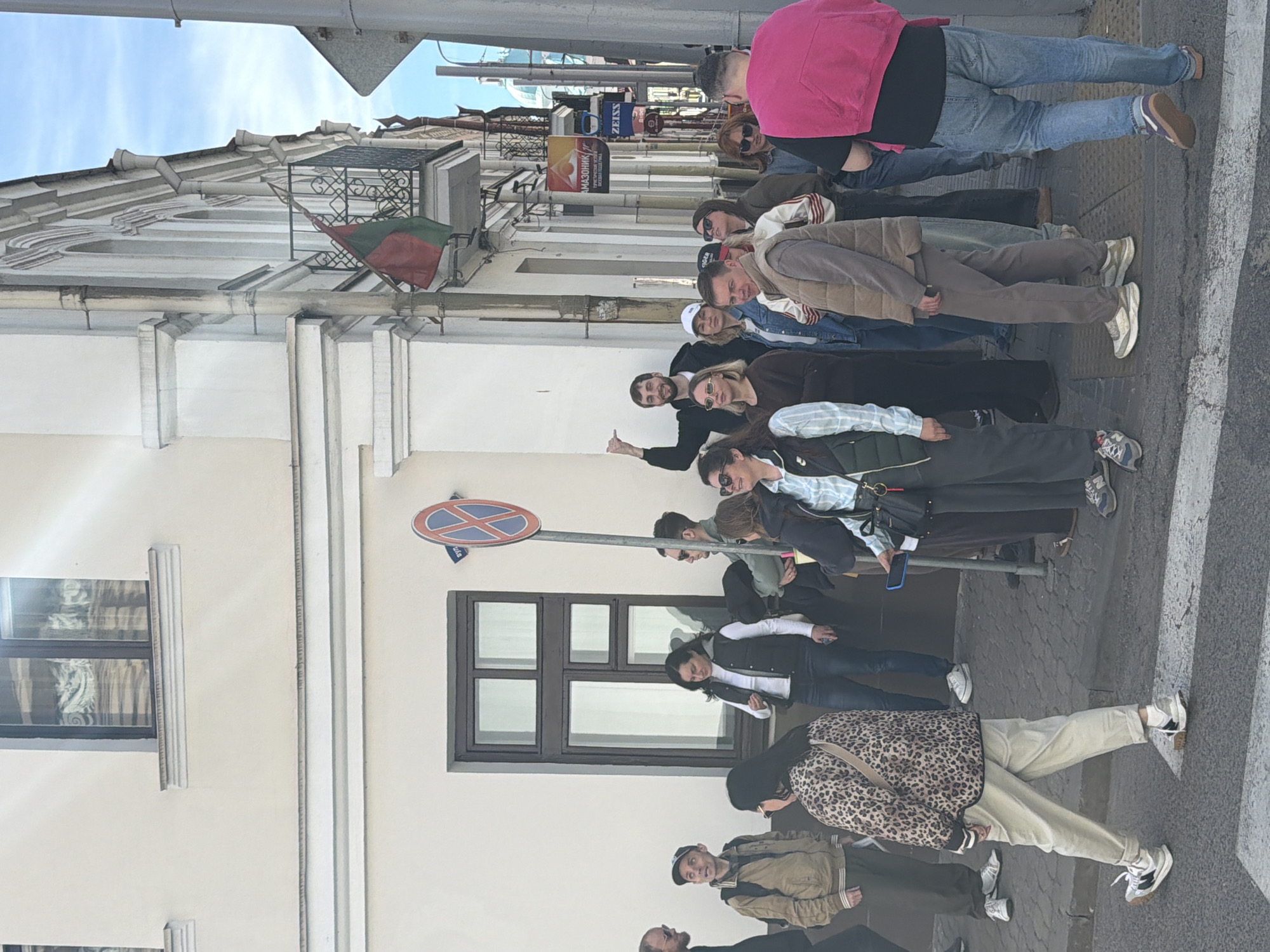 The image captures a group of approximately 20-25 adults gathered on a sidewalk in an urban setting, likely Hrodna, Belarus, during daytime. The weather appears bright and clear, with a blue sky overhead.
The subjects are casually dressed in jackets, hoodies, and long-sleeved shirts, suggesting mild weather. Many are looking towards the camera or slightly off-camera. A man in a black shirt in the middle ground is giving a thumbs-up, indicating a positive or relaxed mood. In the foreground, a woman stands out in a leopard print jacket and light-colored pants.
The scene is set against a backdrop of light-colored, multi-story buildings featuring classical architectural elements like cornices and detailed window frames. An ornate black metal railing adorns a balcony on one building, from which a red and green flag, resembling the Belarusian flag, is draped.
Several commercial signs are visible above the people, including a blue sign displaying "ZEISS" and another with Cyrillic text "МАЗОННИК" (Mazonnik) accompanied by an eye graphic. A prominent red and blue "No Stopping" road sign on a pole stands in the middle ground. White stripes of a crosswalk are visible on the asphalt street surface to the right.
The activity suggests a group outing, possibly a tour or social gathering, as people are clustered together and some are directly engaging with the camera. The overall atmosphere is informal and appears amiable.