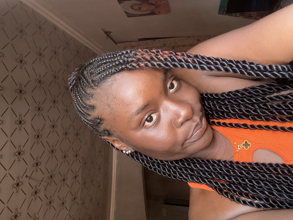 A woman poses, displaying a fresh hairstyle of intricate cornrows leading into thick, two-strand twists. She wears an orange top with a circular cutout, complemented by a gold necklace featuring a clover pendant. Her face is illuminated by natural light, revealing smooth skin and a serene expression. The backdrop showcases a patterned wallpaper, with a framed picture visible in the upper frame. The composition is intimate, focusing on the woman’s features and hairstyle.