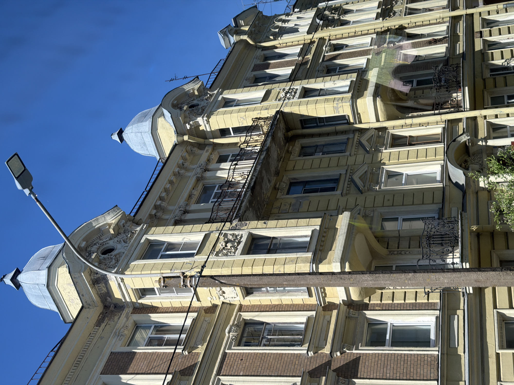 The image shows an upward view of a multi-story building in Kronstadt, Russia. The building features a cream-colored facade with decorative architectural details, including ornate window frames and balcony railings. It has a rooftop with small turrets topped with conical roofs. The sky is blue, and a street lamp is visible to the left. The building’s facade includes brick and masonry elements, and the overall style appears to be historical.