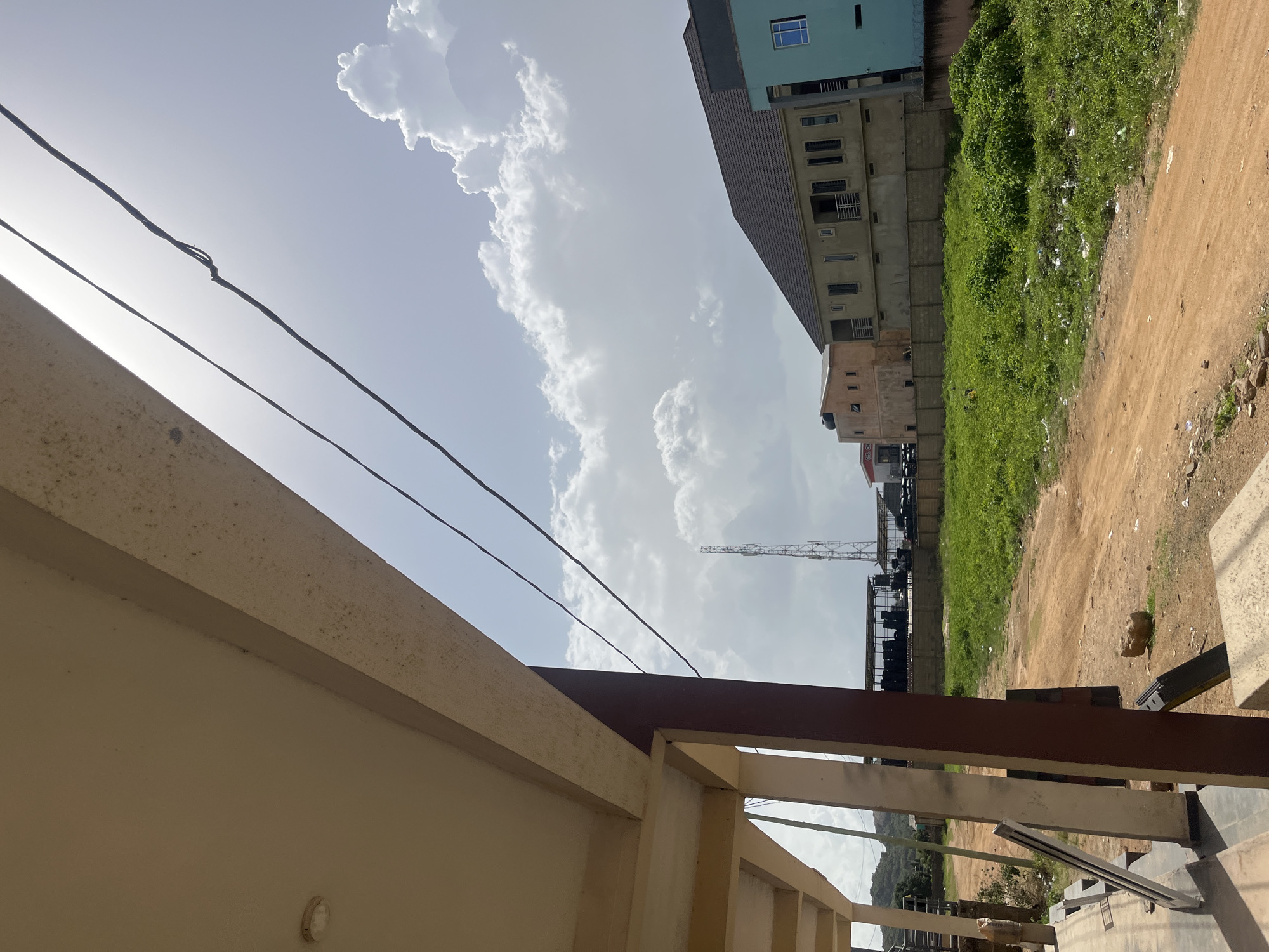The media file depicts an outdoor scene in Gura Top, Nigeria. In the foreground, a cream-colored wall and a dark brown railing suggest a balcony or patio. Several electrical wires are visible, stretching across the frame. Beyond these, the sky is bright with large, white cumulus clouds, indicating daytime. The sun appears to be high, casting a bright light that washes out some of the details.
In the mid-ground, a cluster of buildings of varying heights and colors are visible. One building is painted a light blue and has a grid-like pattern on its facade. Adjacent to it are buildings with a darker, tiled roof and a lighter beige exterior, featuring multiple windows. Further into the background, a tall, lattice-like structure, possibly a transmission tower, rises against the sky.
To the right of the buildings, a patch of lush green vegetation, overgrown with weeds and scattered with debris, meets a dusty, light brown dirt path. The presence of the buildings, the vegetation, and the dirt path suggests an urban or semi-urban environment, possibly under development or with areas of untended land. The overall impression is one of a bright, sunny day in a populated area of Nigeria. No people are clearly visible in the image, and no text is discernible.