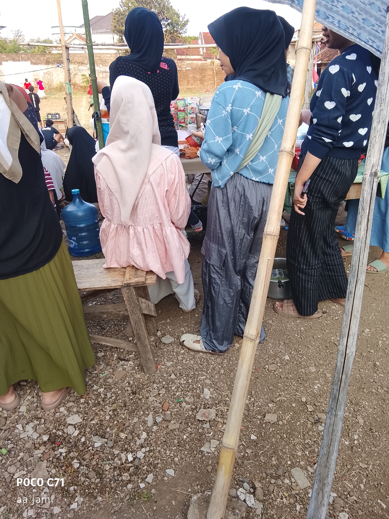 The image shows a group of people, mostly women, standing and sitting outdoors. In the foreground, a woman in a black hijab and a black shirt with white polka dots is visible from the back. To her left, another woman wearing a light beige hijab and a pink and white striped long-sleeved shirt is seated on a wooden bench. Beside her is a large blue water jug. To the right of the woman in the black hijab, a woman in a blue patterned long-sleeved shirt and grey pants is standing. Further to the right, another woman in a black and white striped outfit is visible. The ground is covered in gravel and dirt. In the background, there are buildings and some vegetation. The scene appears to be set in a public outdoor area, possibly a market or a gathering. There is a bamboo pole in the foreground, likely part of a temporary structure. The overall impression is a casual outdoor event or gathering. The image is watermarked with "POCO C71" and "aa jami".