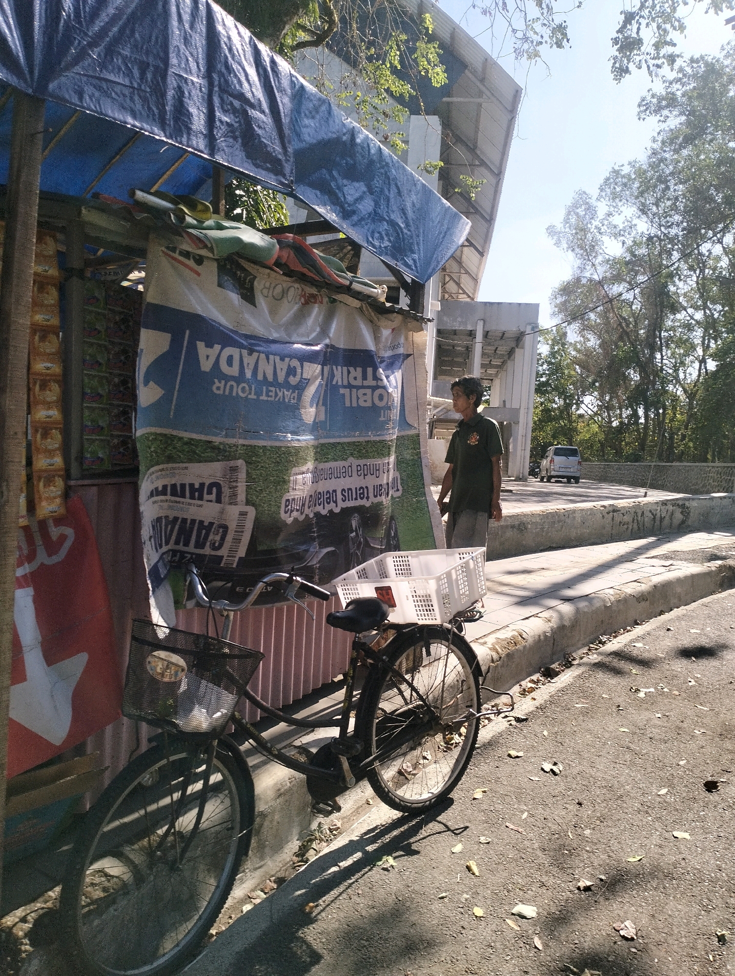 The image depicts a street scene in Purbalingga, Indonesia, on a sunny day. In the foreground, a black bicycle with a white plastic basket on the front is parked by a curb. The bicycle has a black metal frame, two wheels with spokes, and a black seat. To the left of the bicycle is a stall covered with a blue tarp, displaying various items. A large banner on the stall features text in Indonesian, including "PAKET TOUR" and "CANADA". A person, appearing to be male with short dark hair and wearing a dark green t-shirt and grey shorts, stands to the right of the stall, facing away from the camera and looking towards a road in the background. The road is lined with a stone wall and trees. A white minivan is visible driving on the road in the distance. The environment suggests an outdoor market or a roadside stall area in a moderately urban setting, with concrete walkways, asphalt roads, and vegetation.