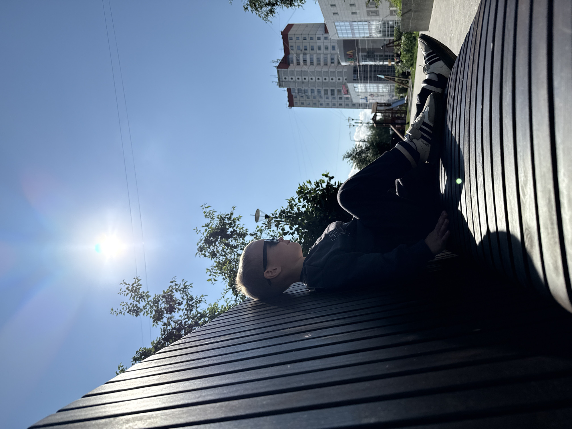 A young person wearing sunglasses and dark clothing, including striped socks and athletic shoes, sits on a dark wooden slatted bench in a park. The sun is bright and low in the sky, casting long shadows. In the background, there are trees, streetlights, a children's playground, and modern apartment buildings. One building appears to be the residential complex "Permskie Vremena" (Perm Times), located in Perm, Russia. The person is seen from the side, looking towards the right of the frame.