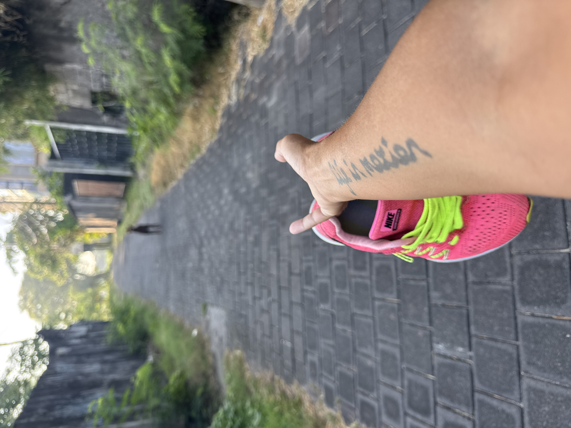 The image shows a first-person perspective of a person holding a pink Nike running shoe with neon green laces in Kuta, Indonesia. The person is standing on a paved road, lined with greenery and buildings. The person's arm is visible, featuring a tattoo on the forearm. The image is slightly blurry, particularly in the background. The shoe is prominently displayed in the foreground.