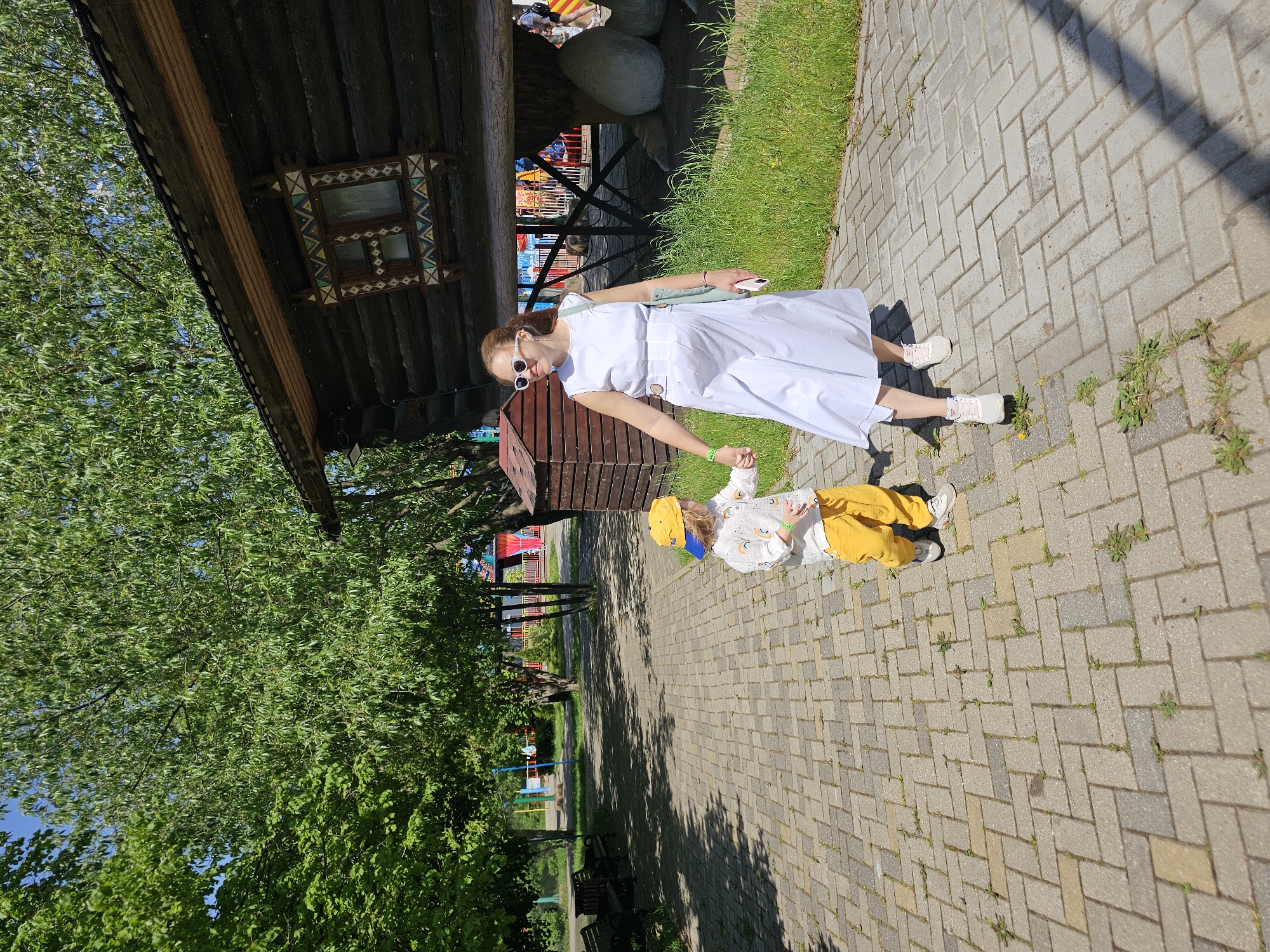 Scene: An outdoor scene in Khimki, Russia, likely in a park or recreational area.
People:
- A woman wearing a white dress, white sunglasses, and carrying a light green bag. She is holding hands with a young child.
- A child wearing a light-colored shirt with rainbow patterns, yellow pants, a yellow hat, and white shoes.
Objects:
- A wooden building with a decorated window frame.
- A small wooden structure resembling a birdhouse.
- Paved walkway with brick patterns.
- Trees with green foliage.
- Green grass.
- Benches along the walkway.
- A building with colorful elements (possibly a playground or amusement park ride) in the background.
- Large, round stones near the wooden building.
Actions:
- The woman is holding the child's hand, presumably walking.
- The child is lying down on the walkway.
Interactions:
- The woman and child are holding hands, suggesting a familial relationship.
Environment:
- Sunny, daytime conditions.
- A park-like setting with trees, grass, and paved walkways.
- A building of possible recreational use visible in the background.