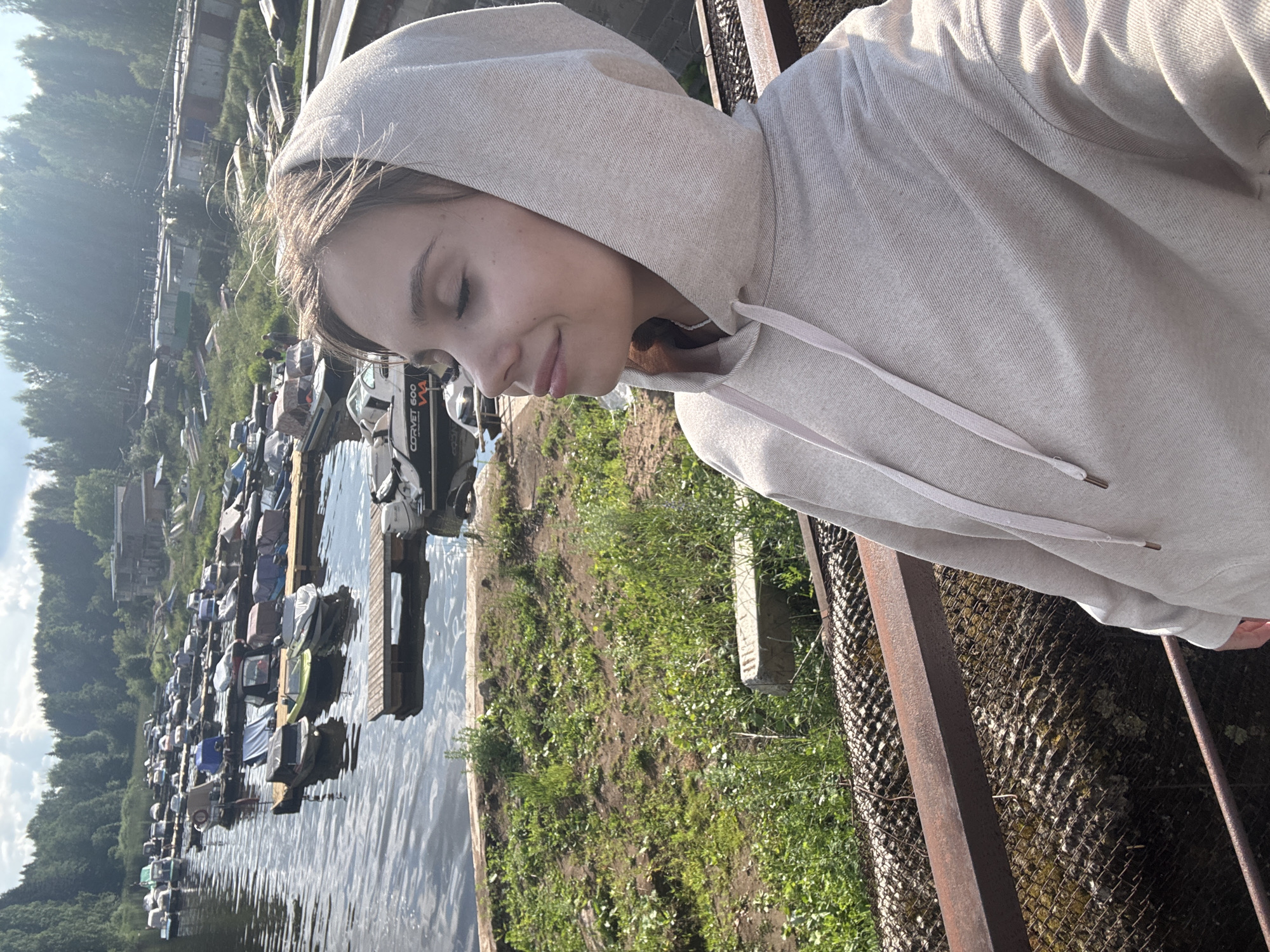 The image depicts a young woman wearing a light-colored hooded sweatshirt, with her eyes closed and a slight smile, enjoying the sunlight. She is standing on a raised platform or walkway, which appears to be made of metal grating and rusty railings.
In the background, there is a body of water, likely a river or canal, lined with numerous boats of various sizes docked along a wooden pier. Beyond the water, there is a densely wooded area with a clear sky overhead. The scene suggests an outdoor recreational area, possibly a marina or boat docking facility. The presence of buildings and structures along the shoreline indicates a populated area.
The image's context is further refined by its location in Chaikovsky, Russia, a city known for its Kama River embankment and recreational facilities. This setting is consistent with the visual elements present in the photograph.