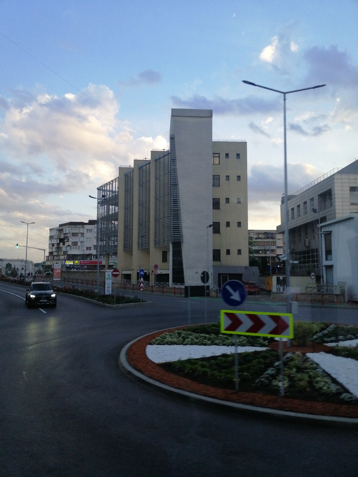 The image depicts a street scene in Fălticeni, Romania, likely during dusk given the lighting. A black car is driving on a wet asphalt road, heading towards the viewer. In the background, several modern buildings of varying heights are visible. One prominent building features a beige facade with a unique angular design and external metal grating. To its right, another building has a light-colored facade and numerous windows. Further in the distance, a lower, multi-story residential building with balconies can be seen. The sky is a soft blue with scattered clouds, and a crescent moon is visible. Streetlights and traffic signs are present, including a blue circular sign with a white arrow pointing left, and a horizontal rectangular sign with alternating red and white chevrons indicating a directional hazard. A roundabout with a landscaped island is also featured in the foreground. The overall impression is of an urban environment with contemporary architecture.