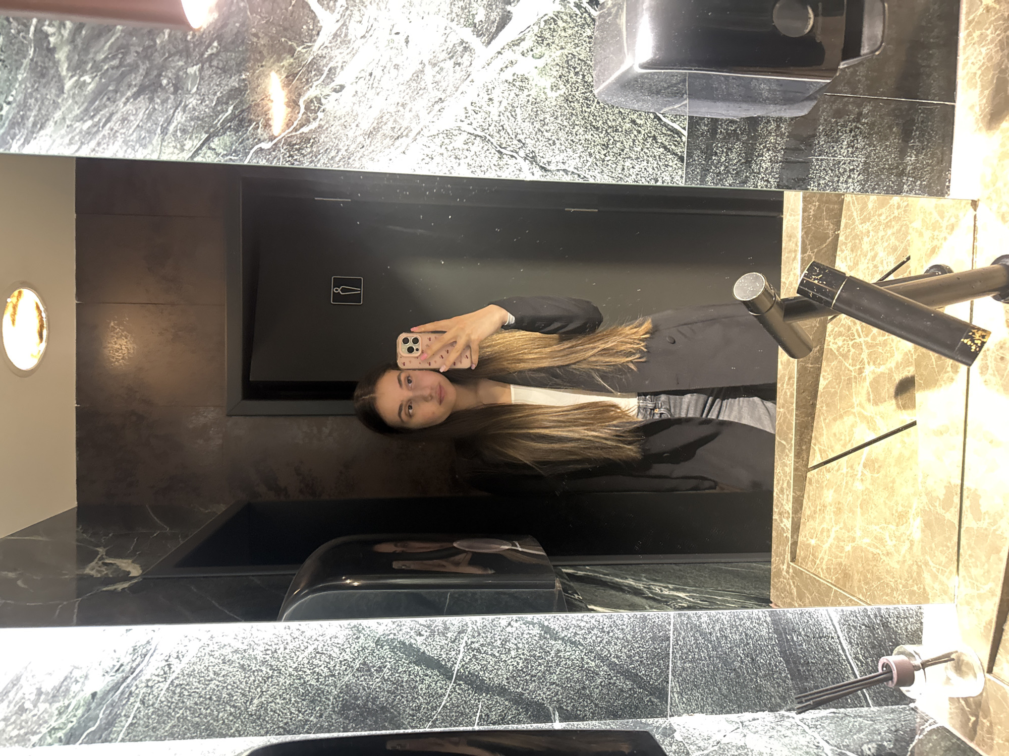A woman is taking a selfie in a bathroom mirror. The bathroom has a dark interior with marble-like surfaces. A hand dryer is visible, as well as modern-style faucets. A door with a restroom sign is behind the woman. The location is possibly in Sofia, Bulgaria, but this is not definitively confirmed by the image.