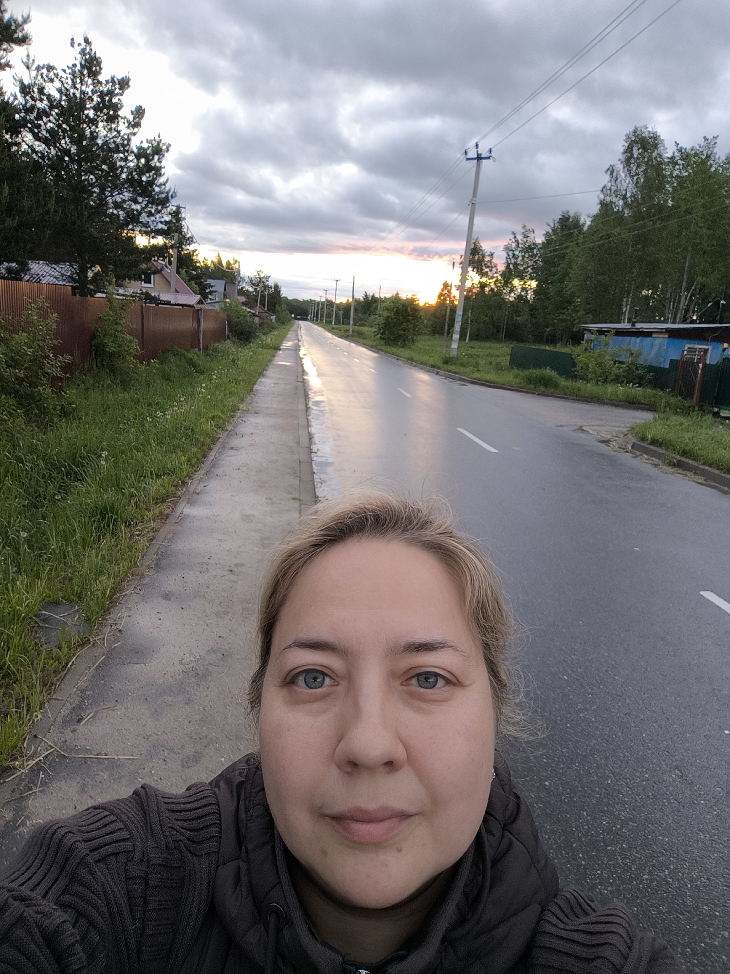 The image captures a woman in the foreground taking a selfie. She has fair skin, light blonde hair, and blue eyes, wearing a dark-colored jacket. Behind her is a wet, asphalt road marked with a white line running through the center. The road appears to lead towards a setting sun or shortly after a rainfall, judging by the reflective surface. On either side of the road, there are grassy areas and foliage, suggesting a rural or suburban environment. To the left, there is a brown fence bordering a residential area. A utility pole with power lines is visible on the right side of the road. The sky is overcast with hints of sunlight breaking through the clouds, indicating either sunrise or sunset. There are trees and vegetation present throughout the scene. It is impossible to verify any location information from the image.