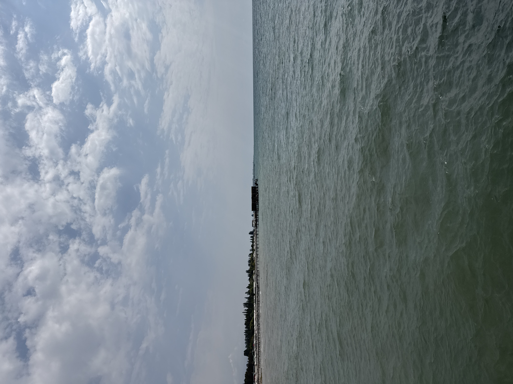 The image depicts a view of the Issyk-Kul lake from the shore in Sary-Oy, Kyrgyzstan. In the foreground, the dark, rippling water of the lake fills the lower half of the frame. In the middle ground, a long wooden pier extends into the water from the shoreline. Along the shore, there is a row of small, white buildings with dark roofs, situated behind a line of trees. A larger, dark building is visible at the far end of the pier. The background consists of a hazy horizon where the lake meets a pale sky filled with scattered white clouds. No people are visible in the image. The scene is calm and depicts a typical lakeside resort setting.