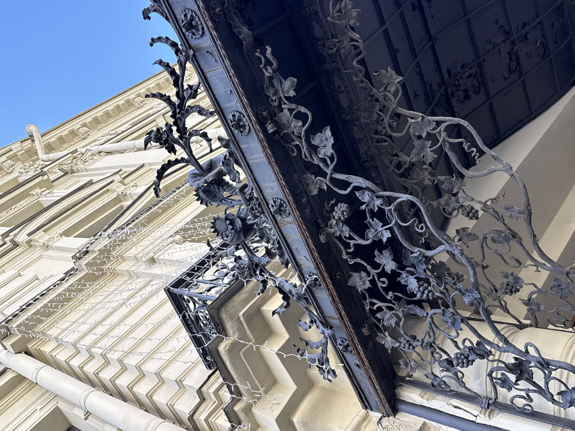 This media file depicts a close-up, low-angle view of an ornate wrought-iron balcony with a vine and grape motif, set against the facade of a building in Saint Petersburg, Russia. The building's architecture features cream-colored walls with detailed decorative cornices and recessed panels, creating a sense of grandeur. A white downspout runs vertically along the building's exterior. White string lights are draped across the facade, adding a festive touch. The sky above is a clear, bright blue, suggesting a sunny day. The perspective emphasizes the intricate craftsmanship of the wrought iron, with its delicate leaves, swirling tendrils, and clusters of grapes. The metalwork appears dark and possibly aged, contrasting with the lighter tones of the building. The overall impression is one of architectural beauty and historical charm, potentially captured during a pleasant time of day, given the clear sky. There is no visible text in the image.
