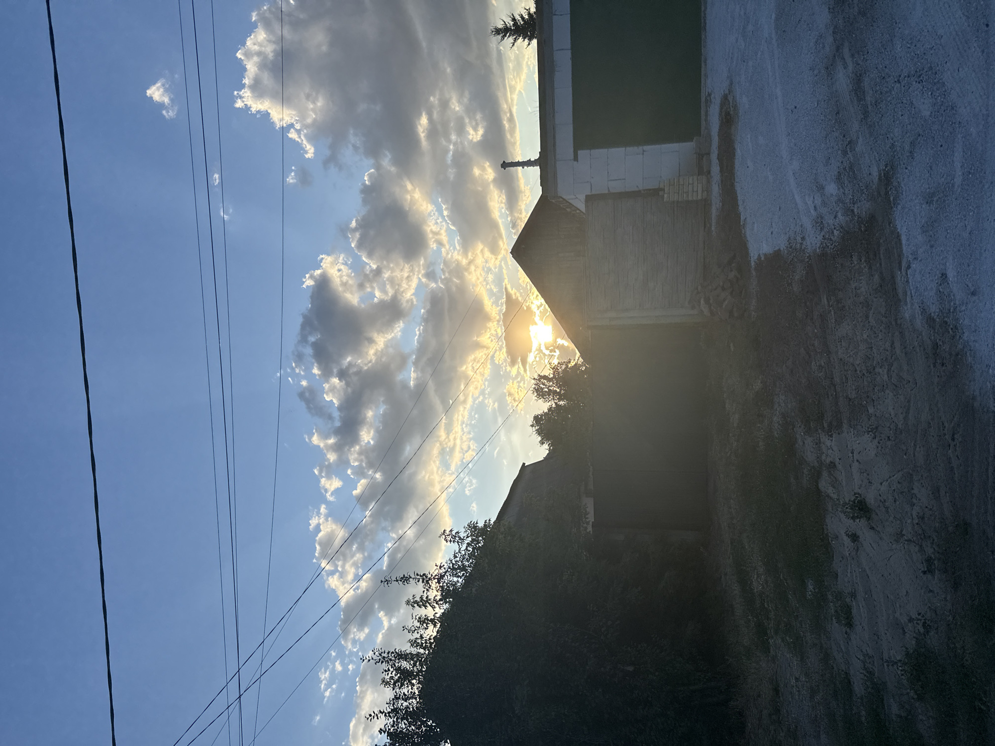 The image depicts a rural or suburban scene at dusk, possibly in Zaporizhzhia, Ukraine, characterized by a dramatic sky with a sun setting behind clouds. In the foreground, there is a gravel or dirt area with sparse vegetation, leading up to structures. On the left, a dense line of trees partially obscures a dark-colored gate and the roofline of a building. To the right, there is a partially constructed building made of white blocks, with a dark garage door. Adjacent to it is a wooden-clad structure with a pitched roof. Overhead, power lines crisscross the sky. The overall lighting suggests late afternoon or early evening. There are no people visible in the image.