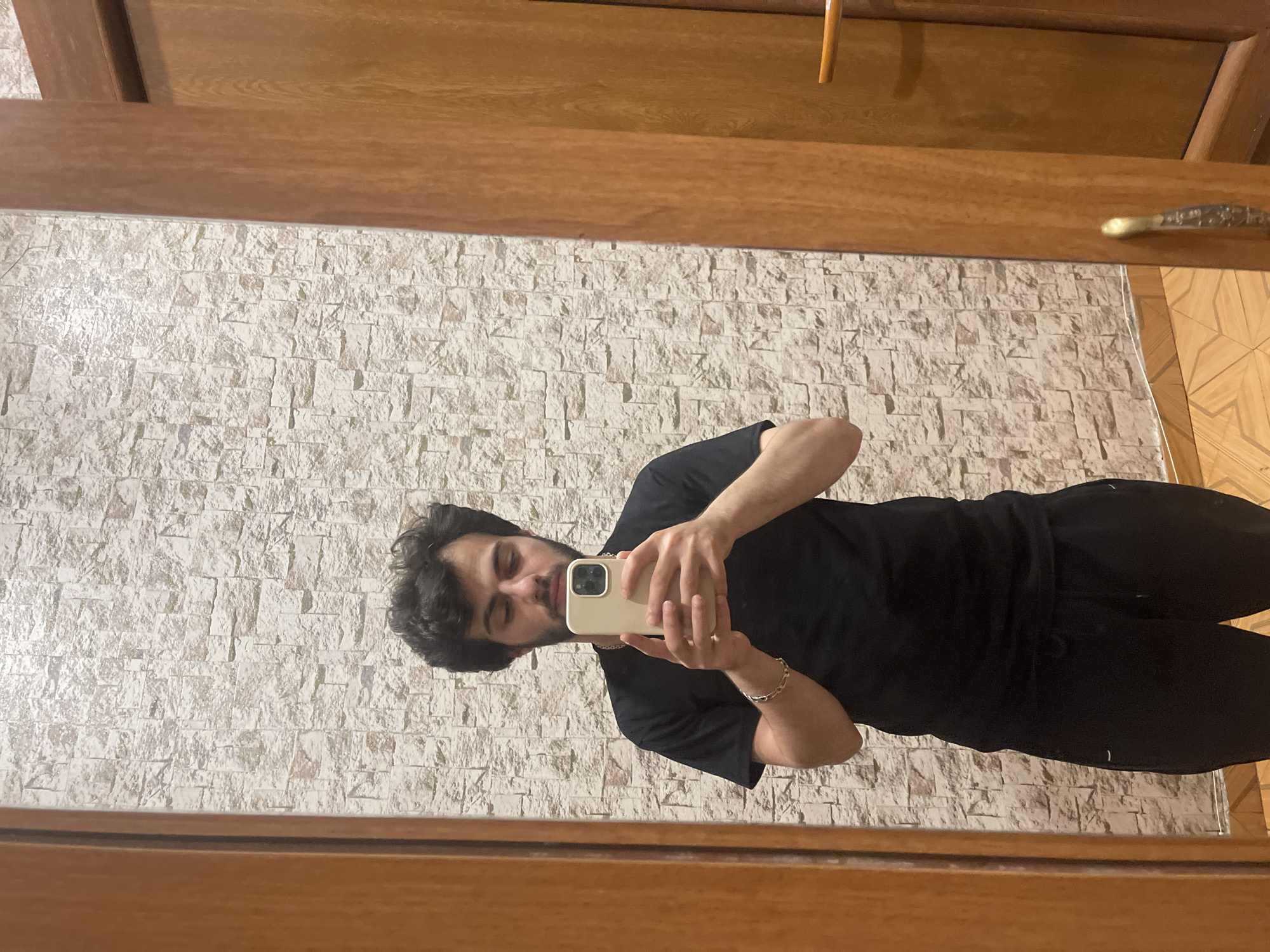 A young man with dark, curly hair and a beard is taking a selfie in a mirror. He is wearing a black t-shirt and black sweatpants. He is holding a light-colored smartphone in his hands, with the screen facing him. A gold chain is visible around his neck, and a gold bracelet is on his left wrist. The background consists of a textured, light-colored wall, possibly wallpaper or stone tiles, and a wooden door frame. The floor is tiled with a wood-grain pattern. The scene appears to be indoors, likely in a residential setting such as a home or apartment in Tbilisi, Georgia, given the context provided. The lighting is artificial and casts a soft glow on the subject and the surroundings.