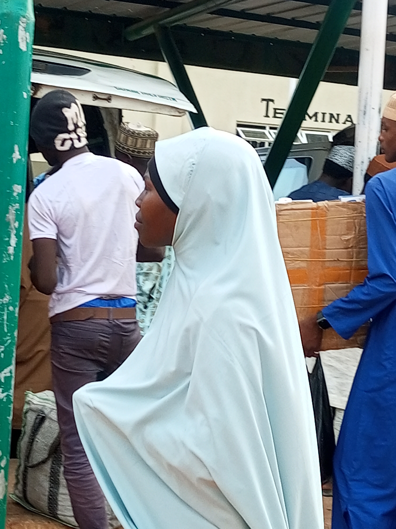 In Gombe, Nigeria, at a terminal, people are gathered near a white vehicle and a structure with a green metal support. A man wearing a black beanie with white lettering is visible in profile, facing left. He is wearing a light pink t-shirt and dark pants, with blue underwear showing above his waistband. Next to him, another man is partially visible, wearing a white and gold patterned cap. In the foreground, a person wearing a light blue hijab is seen from the side, facing right. To the right of the hijabed person, a person in a blue robe is reaching towards a cardboard box. Bags are visible on the ground. The word "TERMINA" is visible on a building in the background.